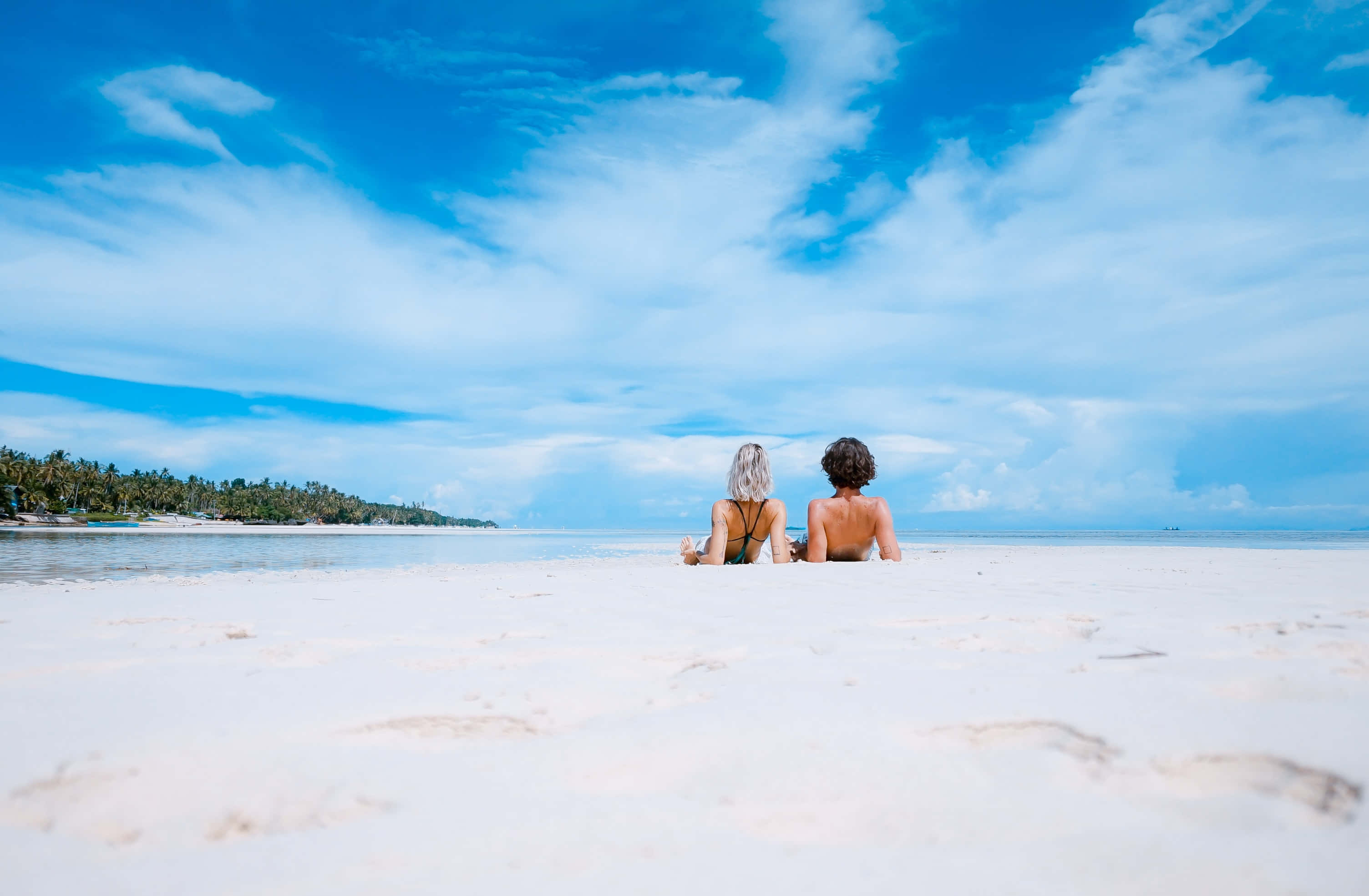 Summer Vacation 桌布 壁紙 — 兩個人並肩坐在純淨的白色沙灘上，面對著碧綠色的海洋，頭頂是廣闊的藍天，點綴著縷縷薄雲。