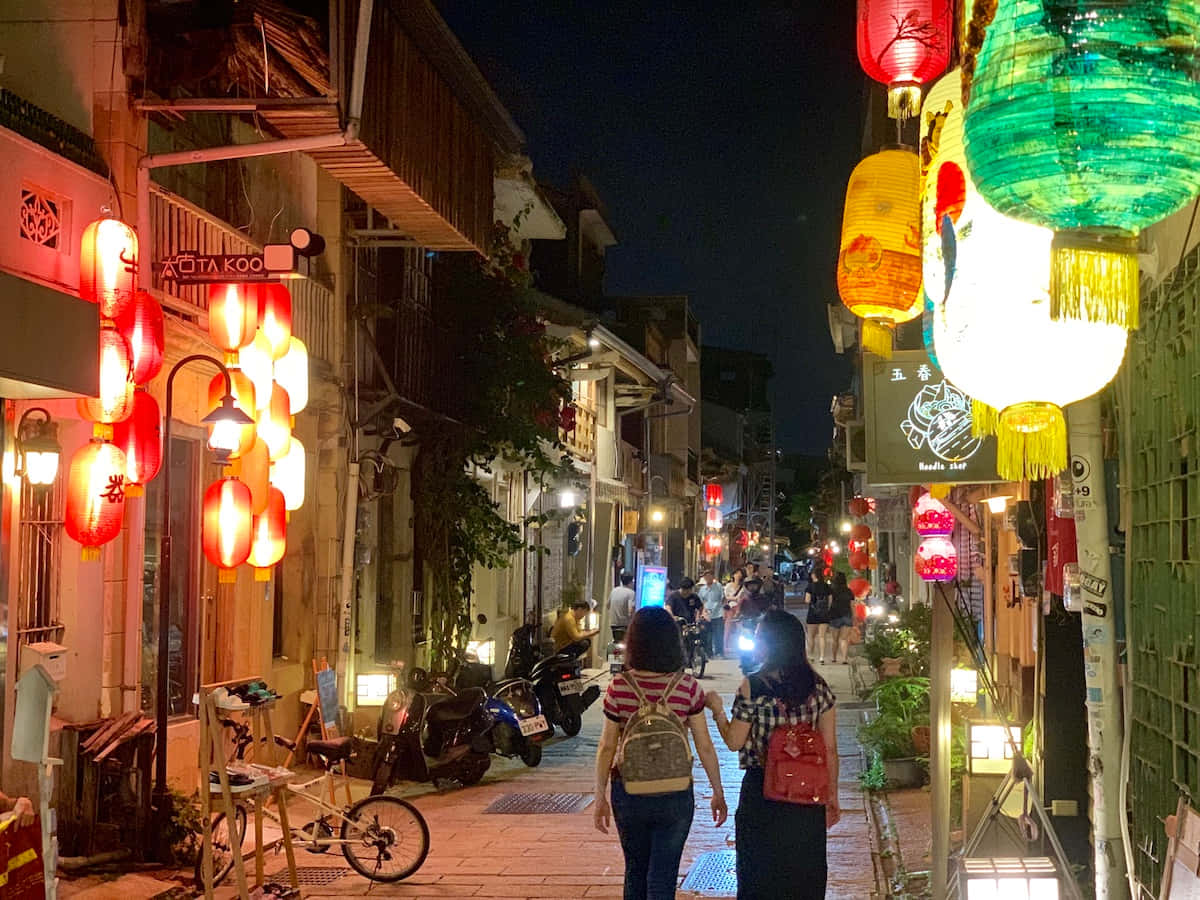 街頭 桌布 壁紙 — 兩個人在一條狹窄的夜晚街道上漫步，兩旁掛著發光的紅色和綠色燈籠，還有傳統建築物。