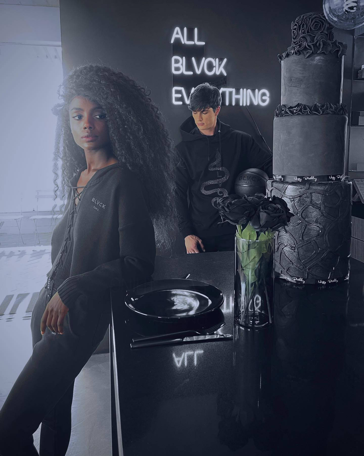 Blvck Paris 桌布 壁紙 — 兩個人在單色房間內，有霓虹燈牌和一個黑色蛋糕放在光面桌上。
