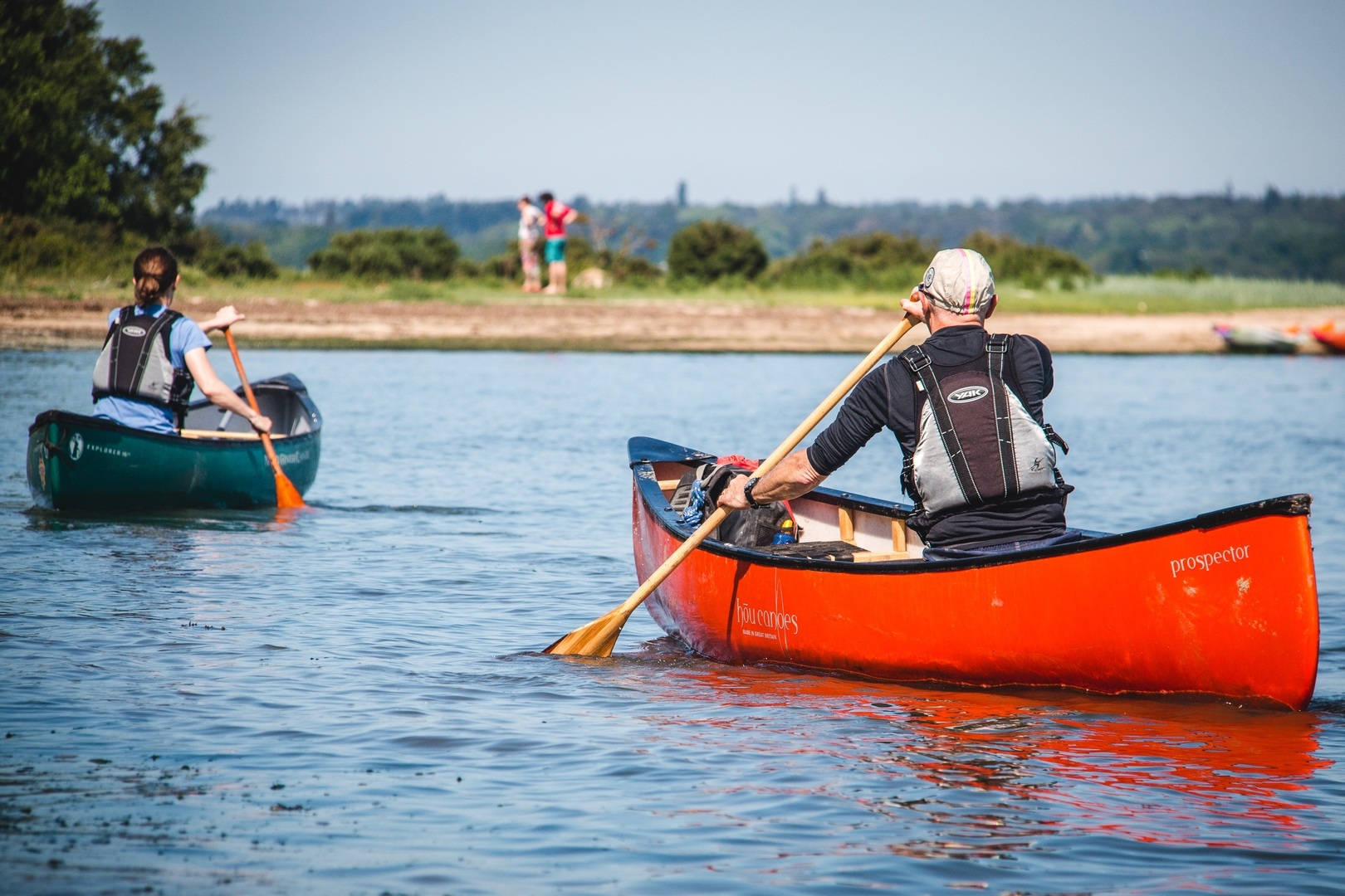 Rowing 桌布 壁紙 — 兩個人在平靜的藍色水面上划著鮮紅和鮮綠的獨木舟，背景有樹木成蔭的岸邊和遠處的人影。