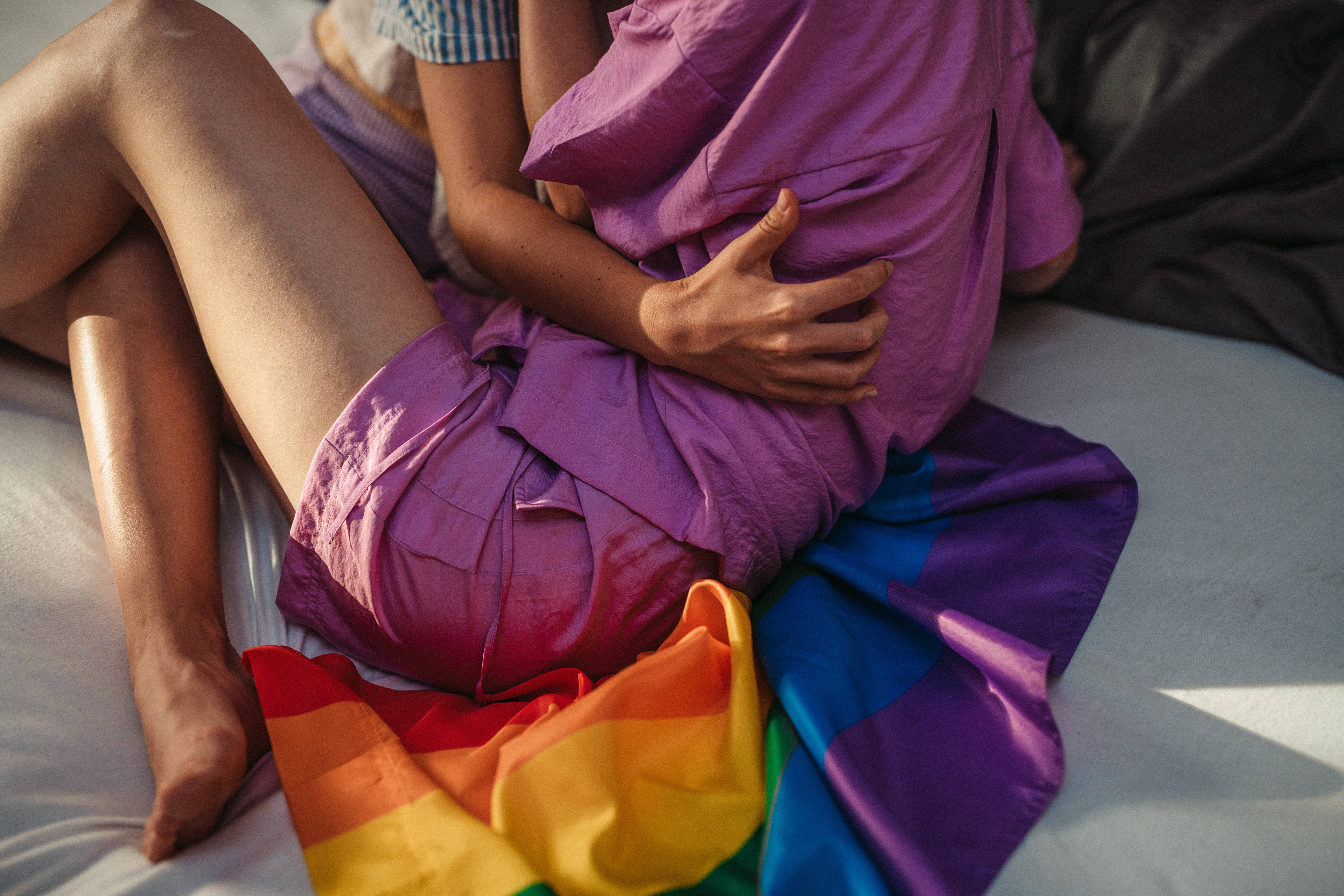 Pride Flag 桌布 壁紙 — 兩個人在床上擁抱，旁邊掛著一面鮮豔的彩虹旗，柔和自然光線下。