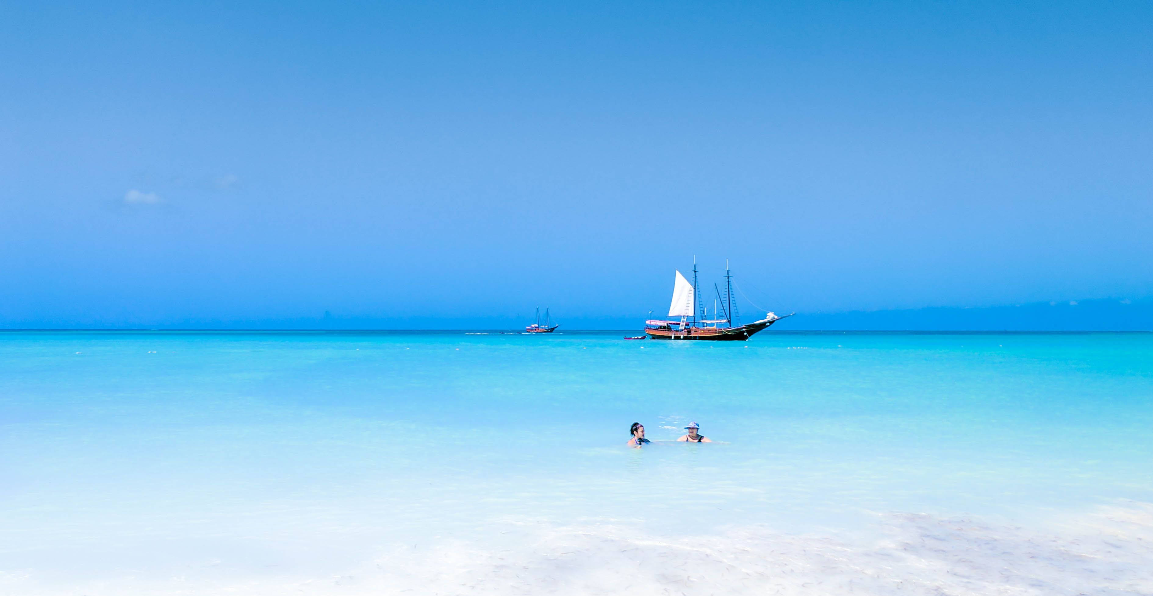Aruba 桌布 壁紙 — 兩個人在廣闊藍天下，於清澈碧綠的海水中靠近傳統帆船游泳。