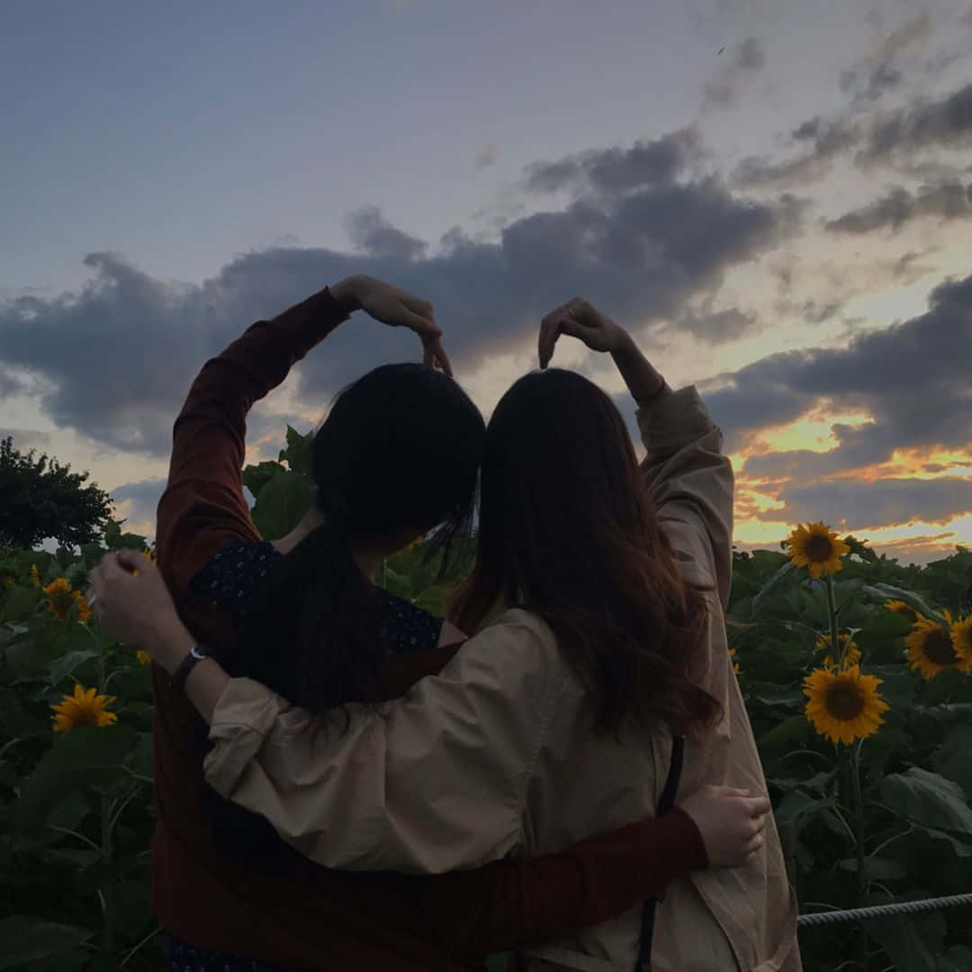 Best Friend Aesthetic 桌布 壁紙 — 兩個人在日落時分的向日葵田裡擁抱，用手比出心形，背景是陰沉的天空。