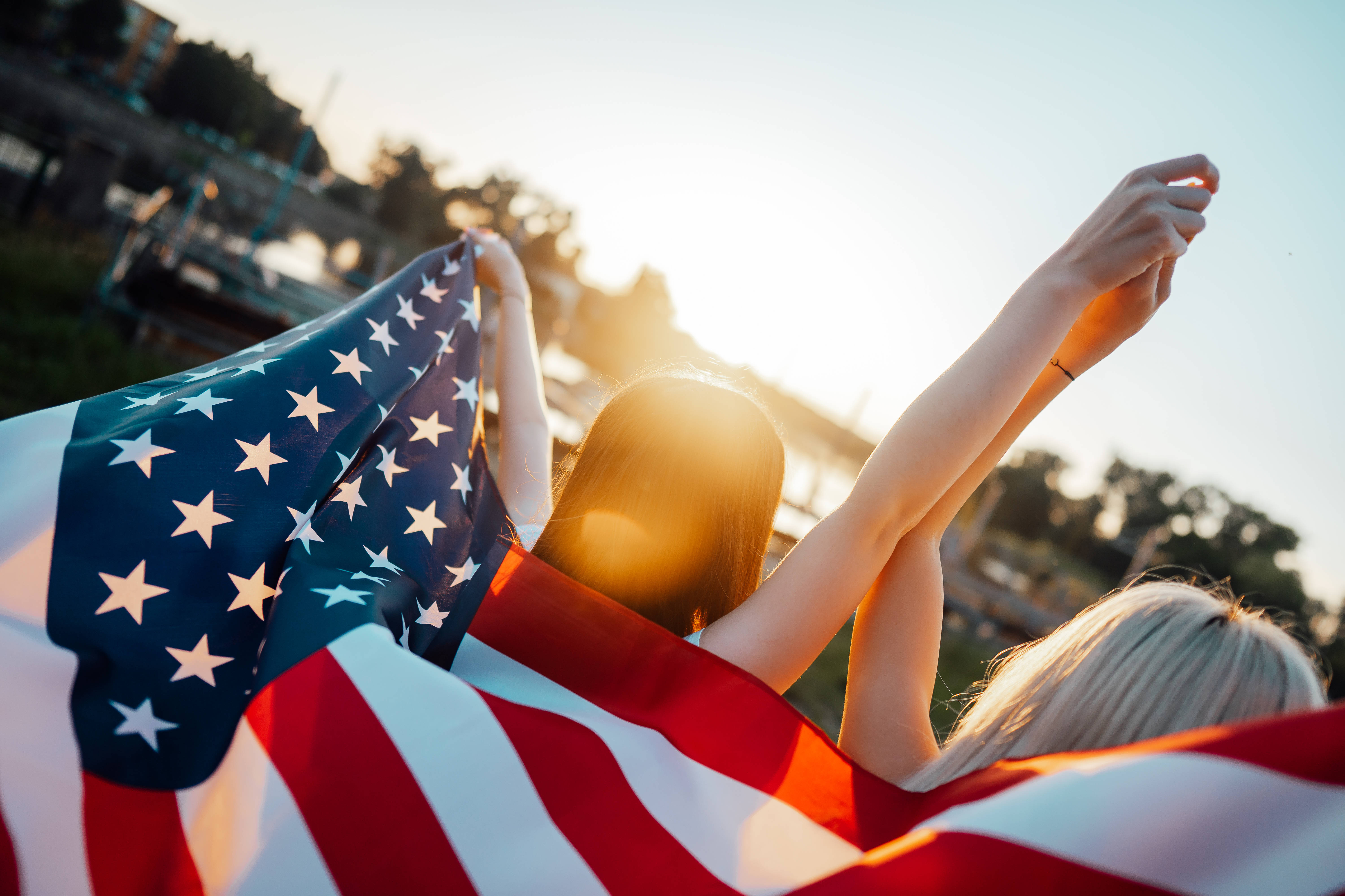 USA flag iPhone 桌布 壁紙 — 兩個人在日落時分高舉雙臂擁抱美國國旗，享受歡樂的戶外時刻。