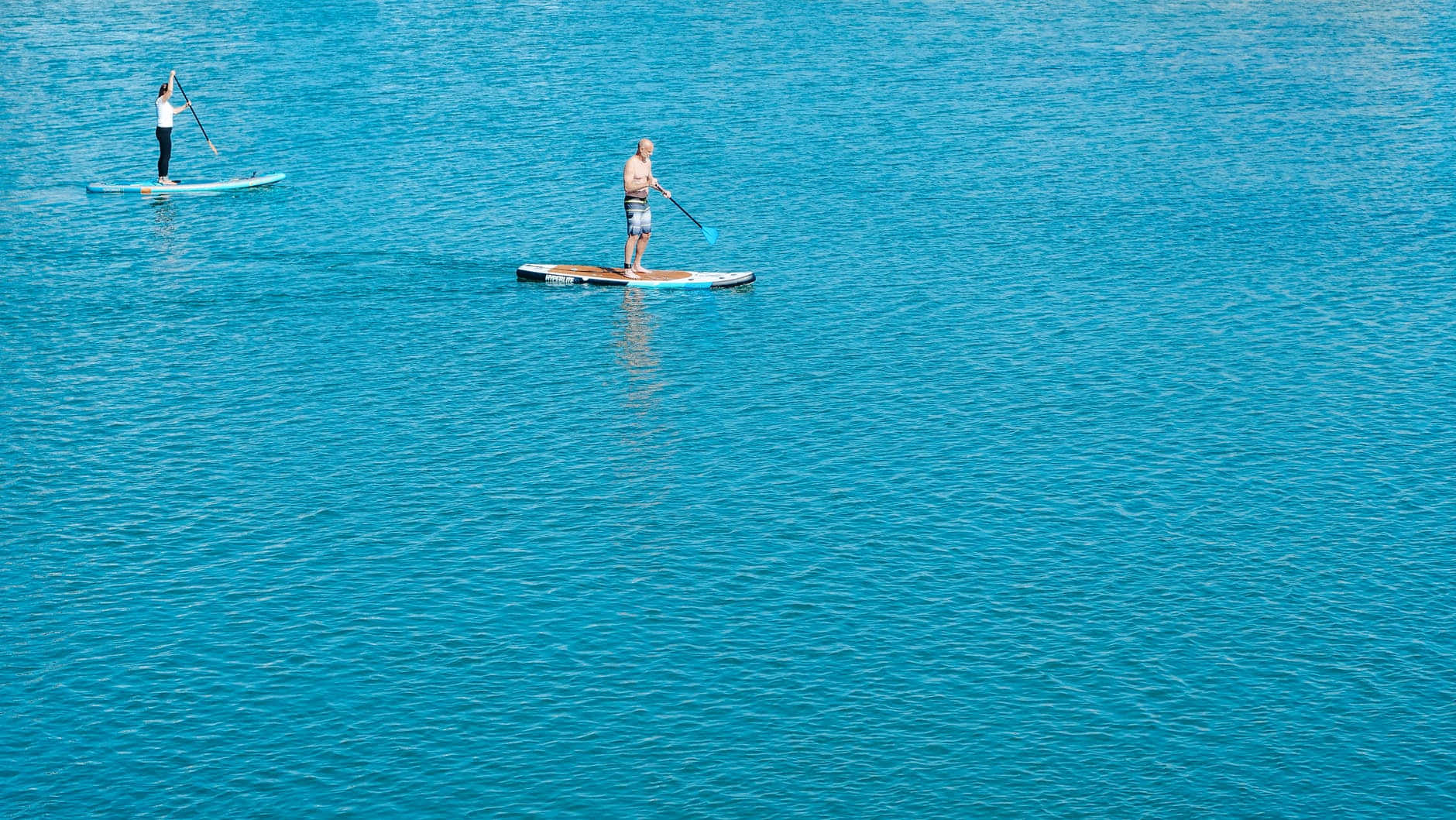 Paddleboarding 桌布 壁紙 — 兩個人在明亮的日光下，在廣闊的碧綠水面上划槳板。