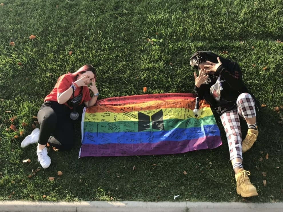 Flag 桌布 壁紙 — 兩個人在明亮的日光下坐在草地上，旁邊是一面佈滿簽名的彩虹旗。