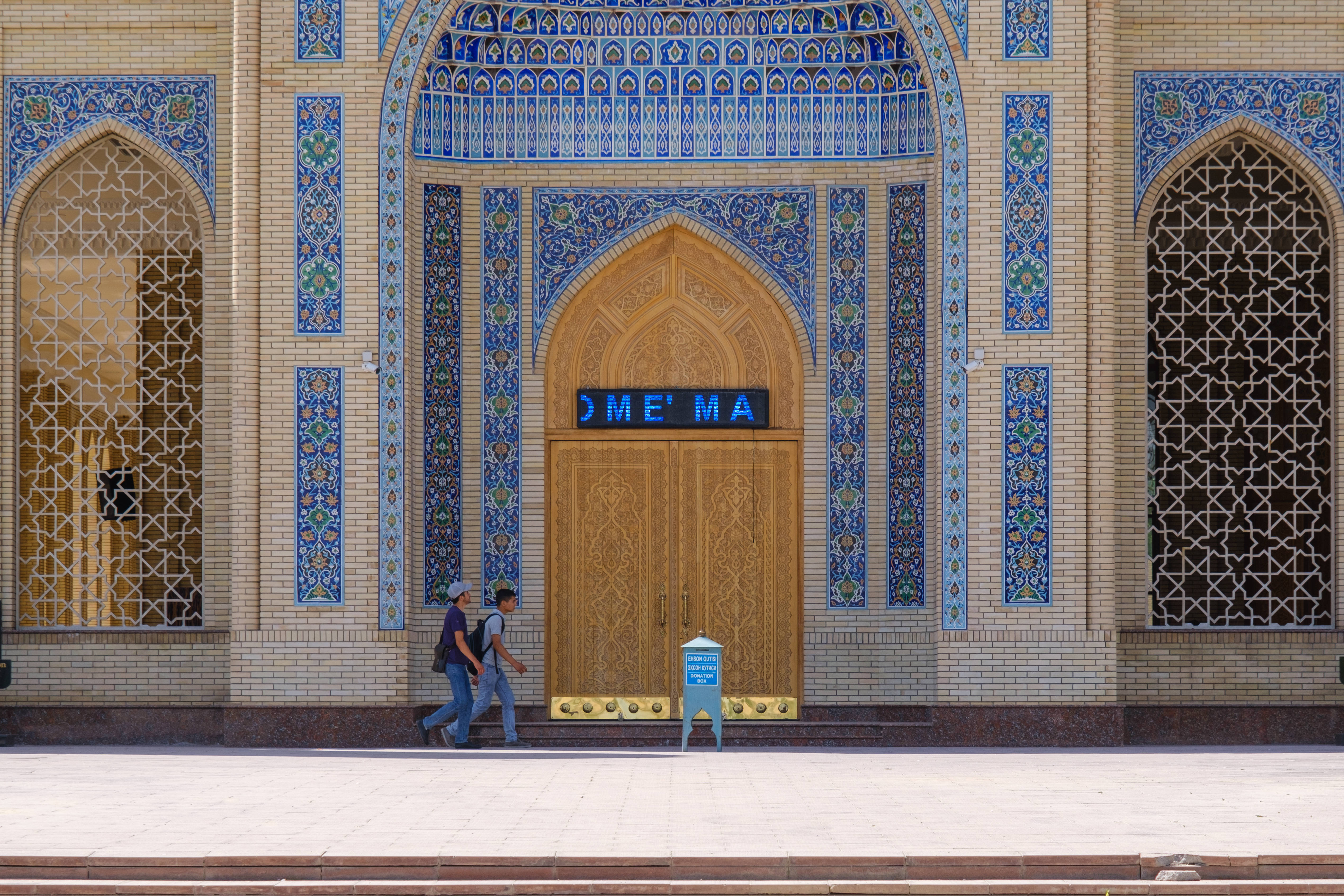 Uzbekistan 桌布 壁紙 — 兩個人在明亮的日光下走過一座宏偉的清真寺入口，入口有精緻的藍色瓷磚和木門。