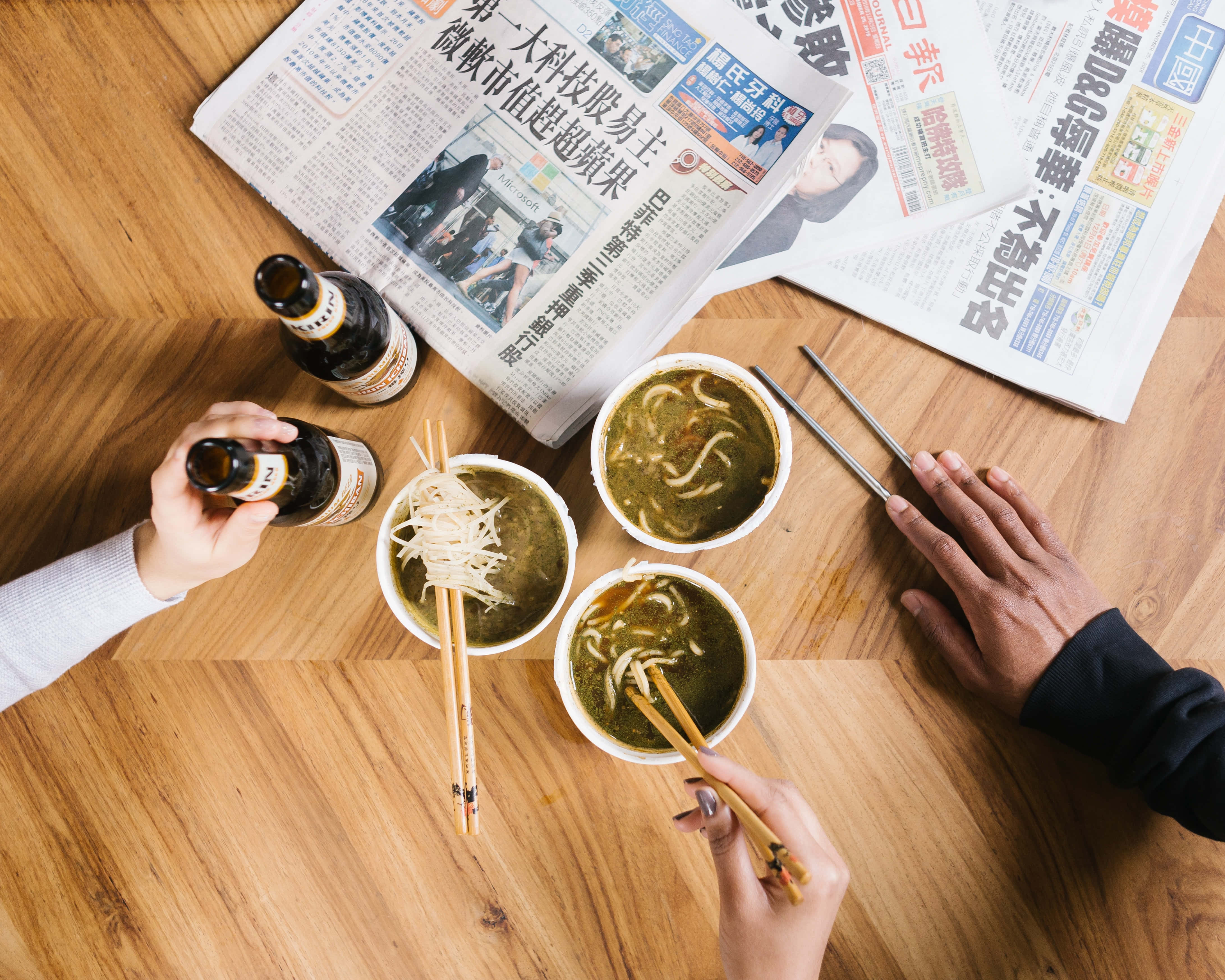 高清 桌布 壁紙 — 兩個人在木桌上享用碗裝湯麵和啤酒，同時閱讀中文報紙。