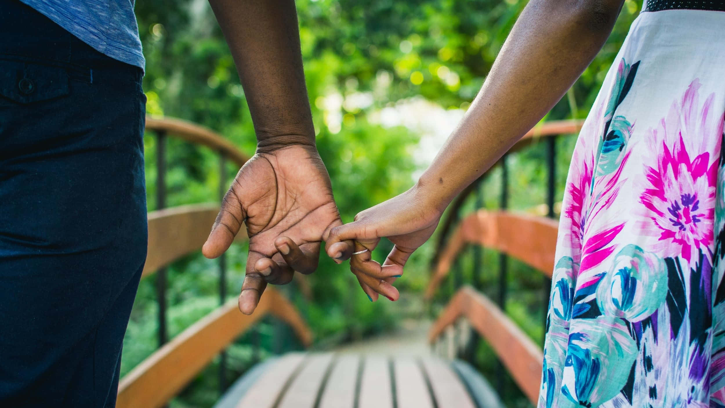 Holding Hands 桌布 壁紙 — 兩個人在木橋上牽手，周圍是茂密的綠色樹葉。