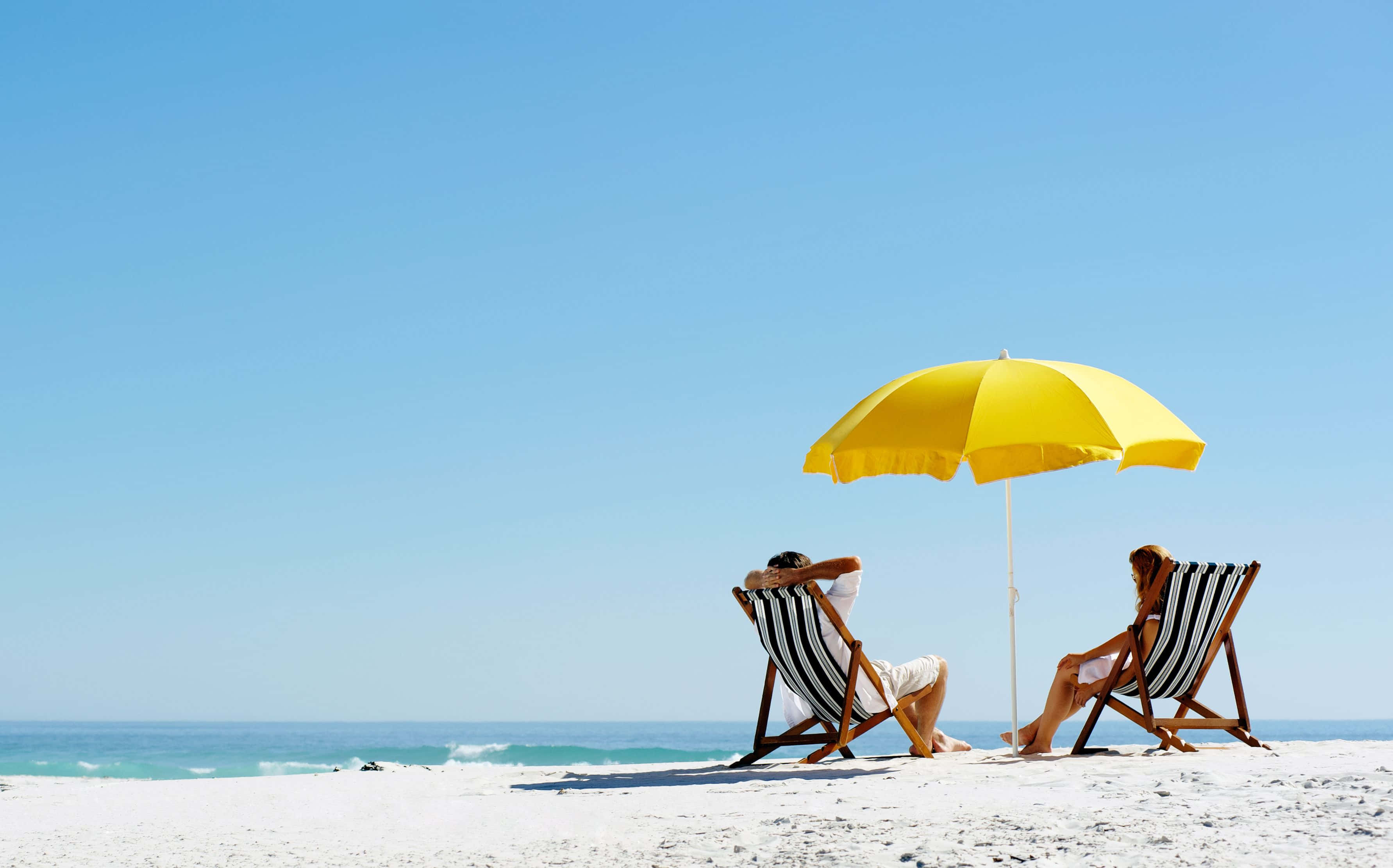 Beach Umbrella 桌布 壁紙 — 兩個人在條紋躺椅上放鬆，在明亮的黃色傘下，面對海洋的原始白色沙灘上。