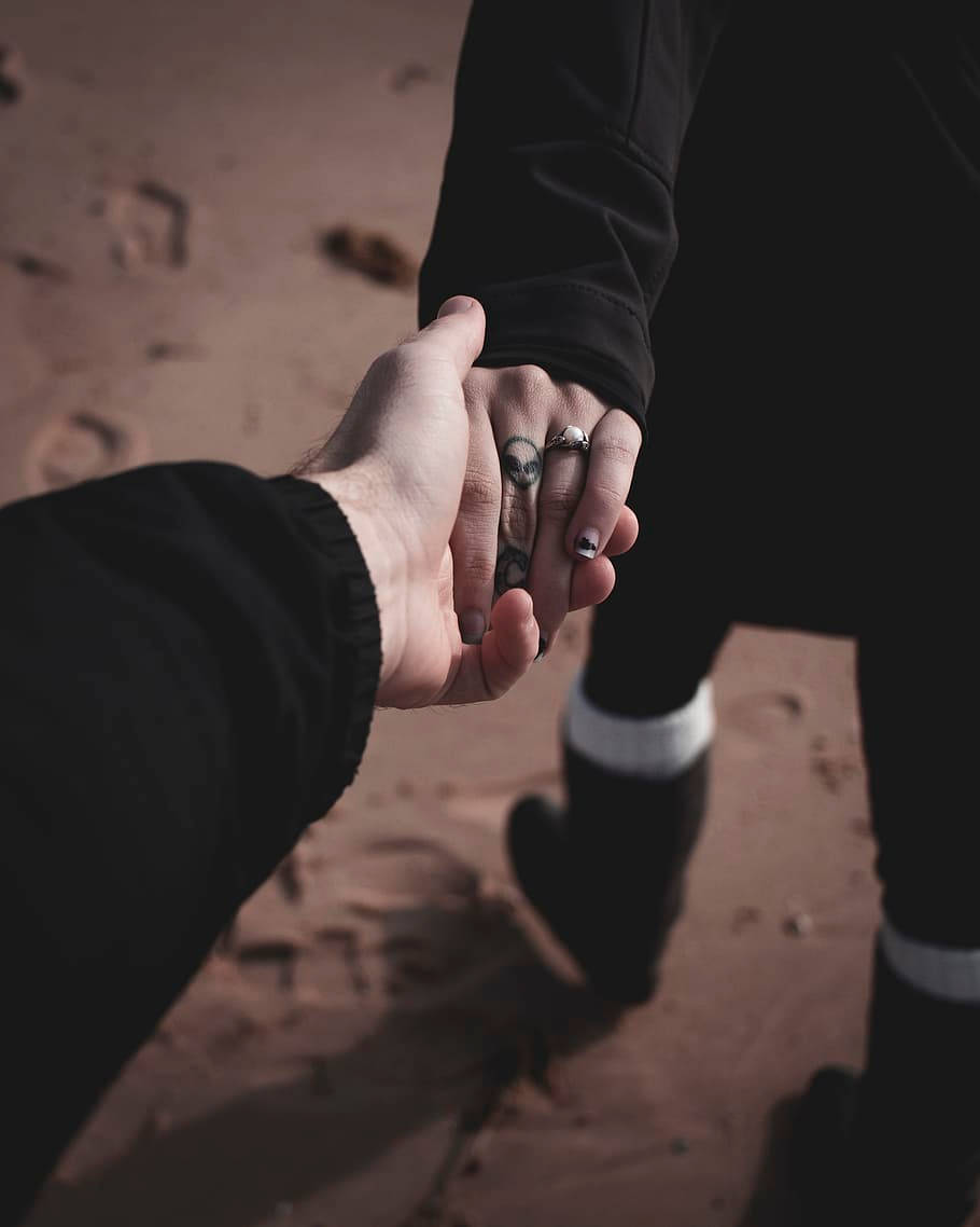 hands 桌布 壁紙 — 兩個人在沙地上牽手，一隻手有刺青和戒指，另一隻手是裸露的，兩人都穿著黑色衣服。