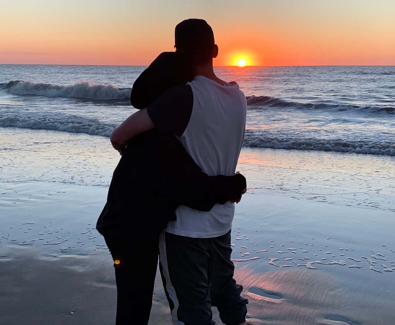 海灘 桌布 壁紙 — 兩個人在沙灘上擁抱，夕陽在海洋上空落下，投射出溫暖的橙色光芒。