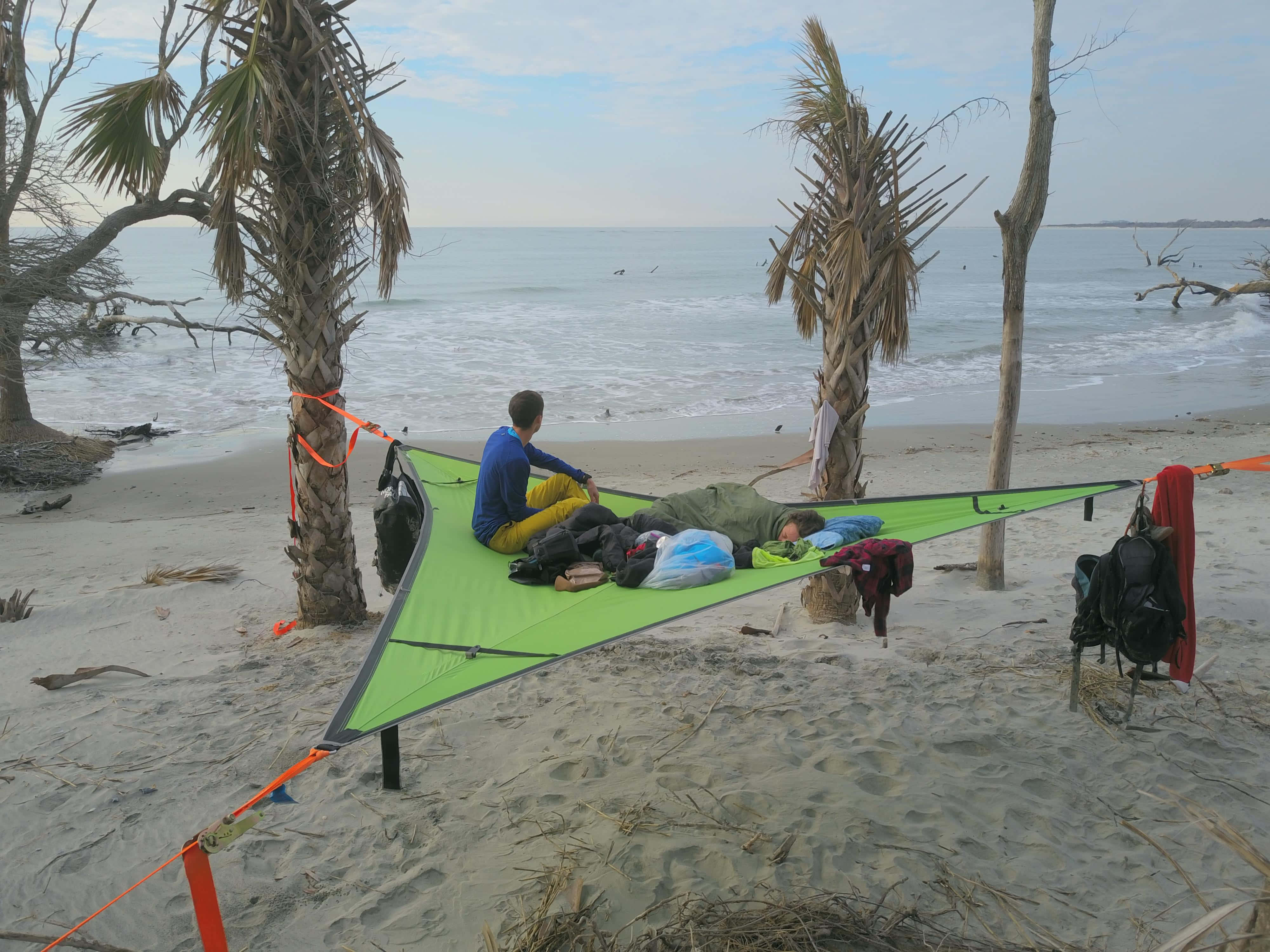 Beach Camping 桌布 壁紙 — 兩個人在沙灘上棕櫚樹之間拉起的亮綠色防水布上放鬆，海浪翻滾而來。