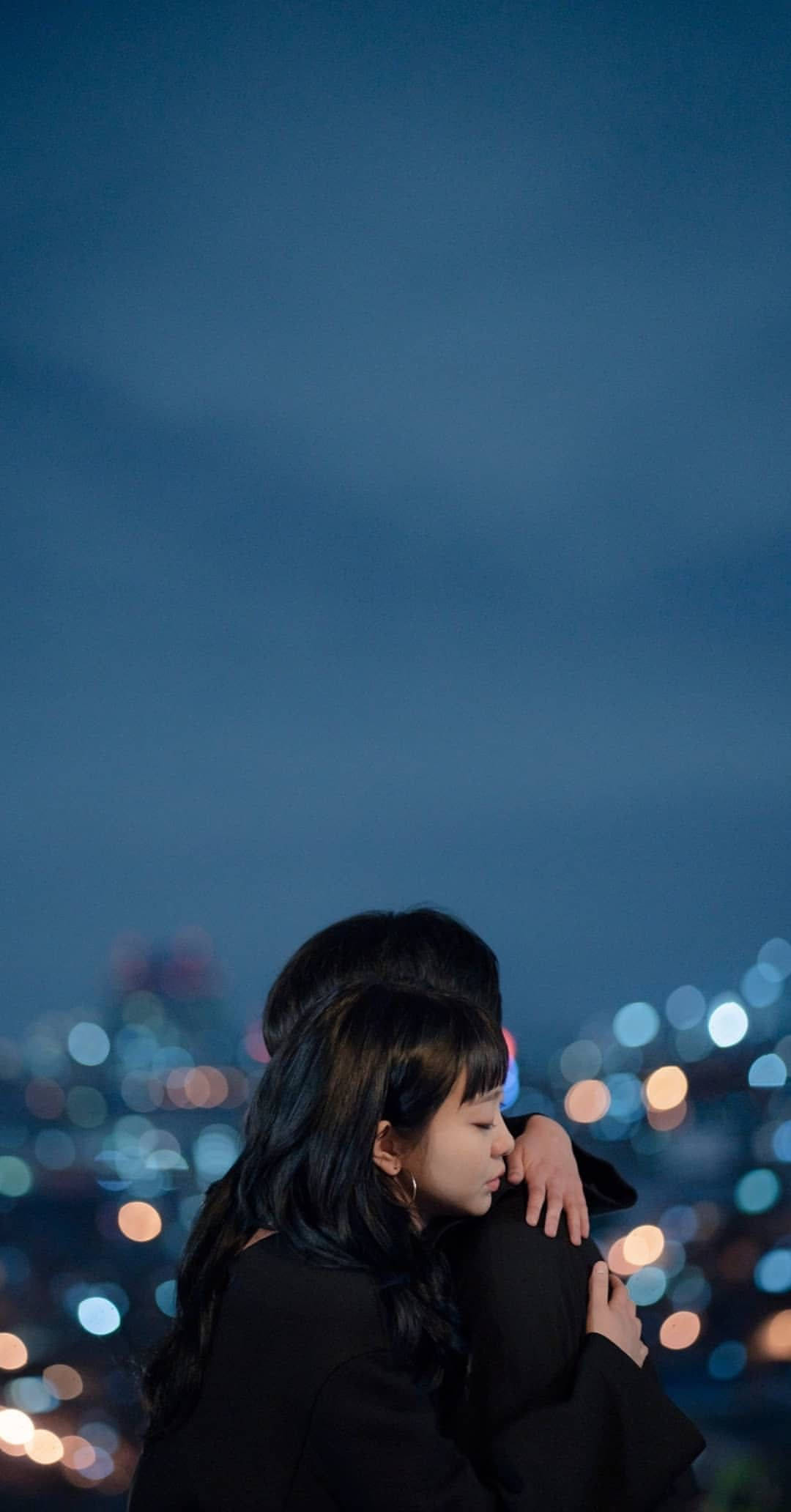 drama 桌布 壁紙 — 兩個人在深藍色暮色天空下，以模糊的城市天際線為背景，溫柔地擁抱，天際線閃爍著散景燈光。