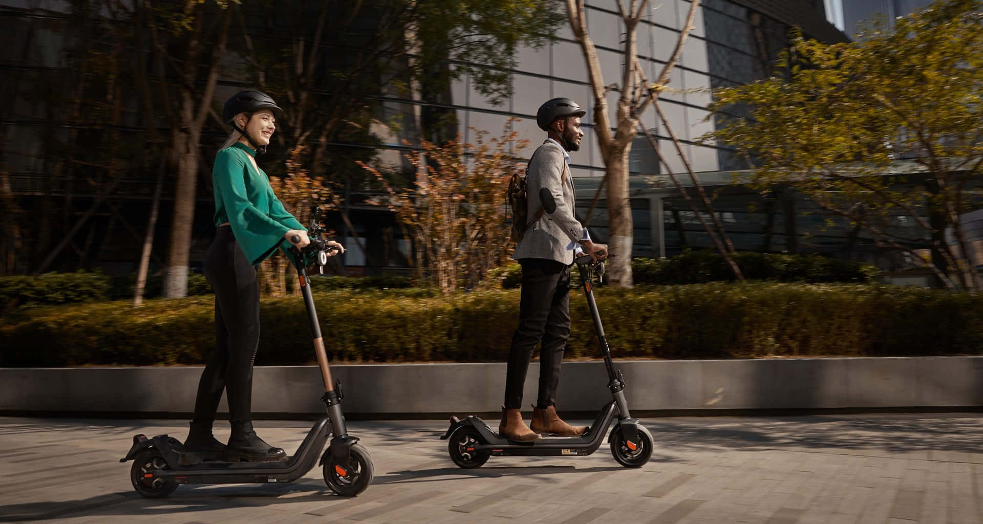 scooter 桌布 壁紙 — 兩個人在溫暖的陽光下沿著一條鋪砌的城市小徑騎電動滑板車，小徑兩旁種滿樹木和現代建築。