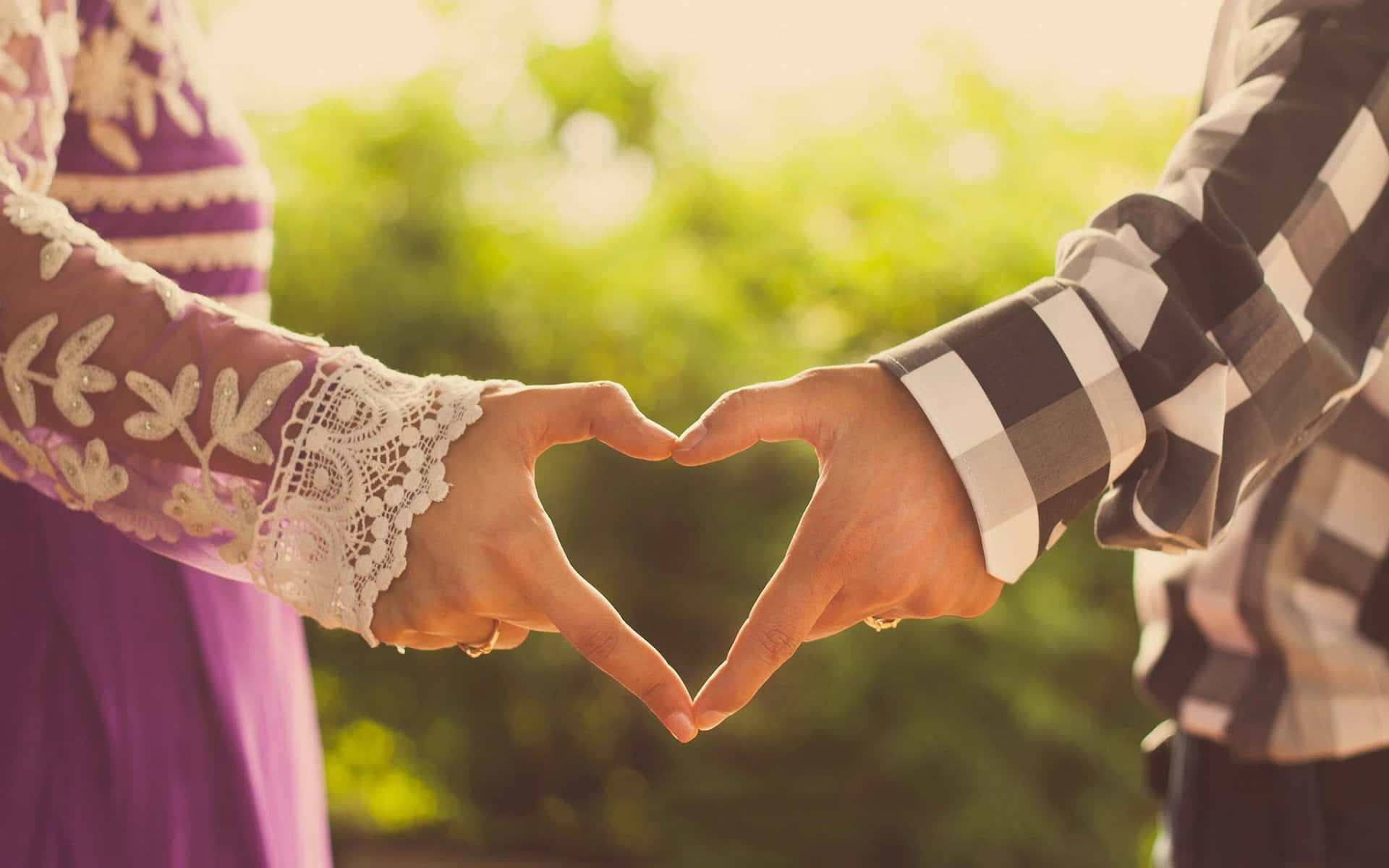 Love Couple 桌布 壁紙 — 兩個人在溫暖陽光沐浴下的柔和綠色模糊背景前，用手形成心形。