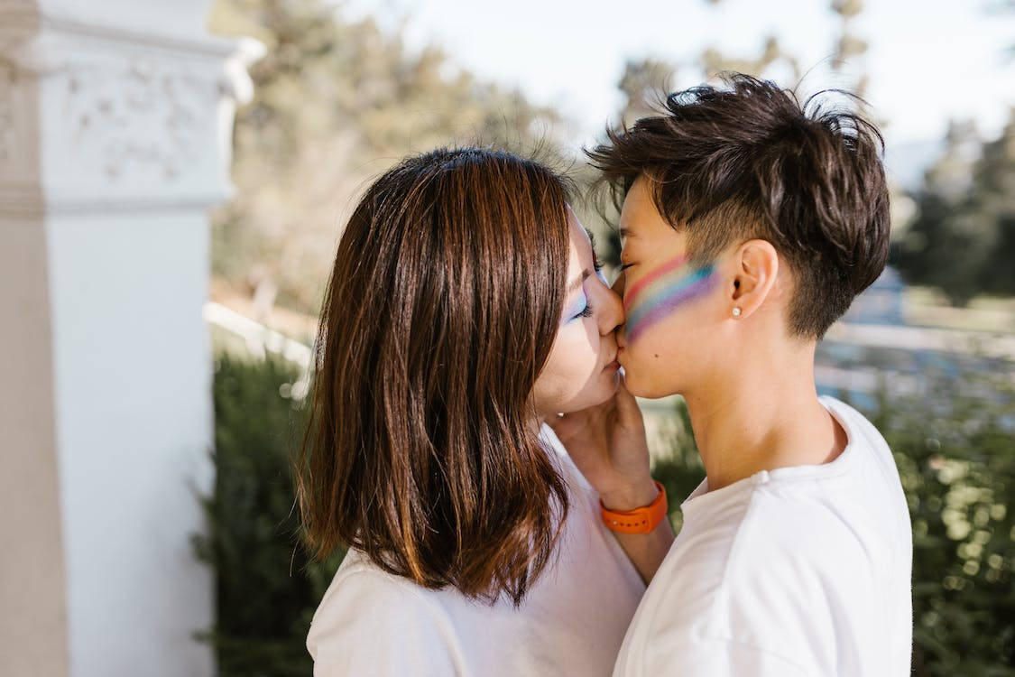 Kissing 桌布 壁紙 — 兩個人在白色柱子旁，在明亮的陽光下，帶著彩虹臉部彩繪在戶外接吻。