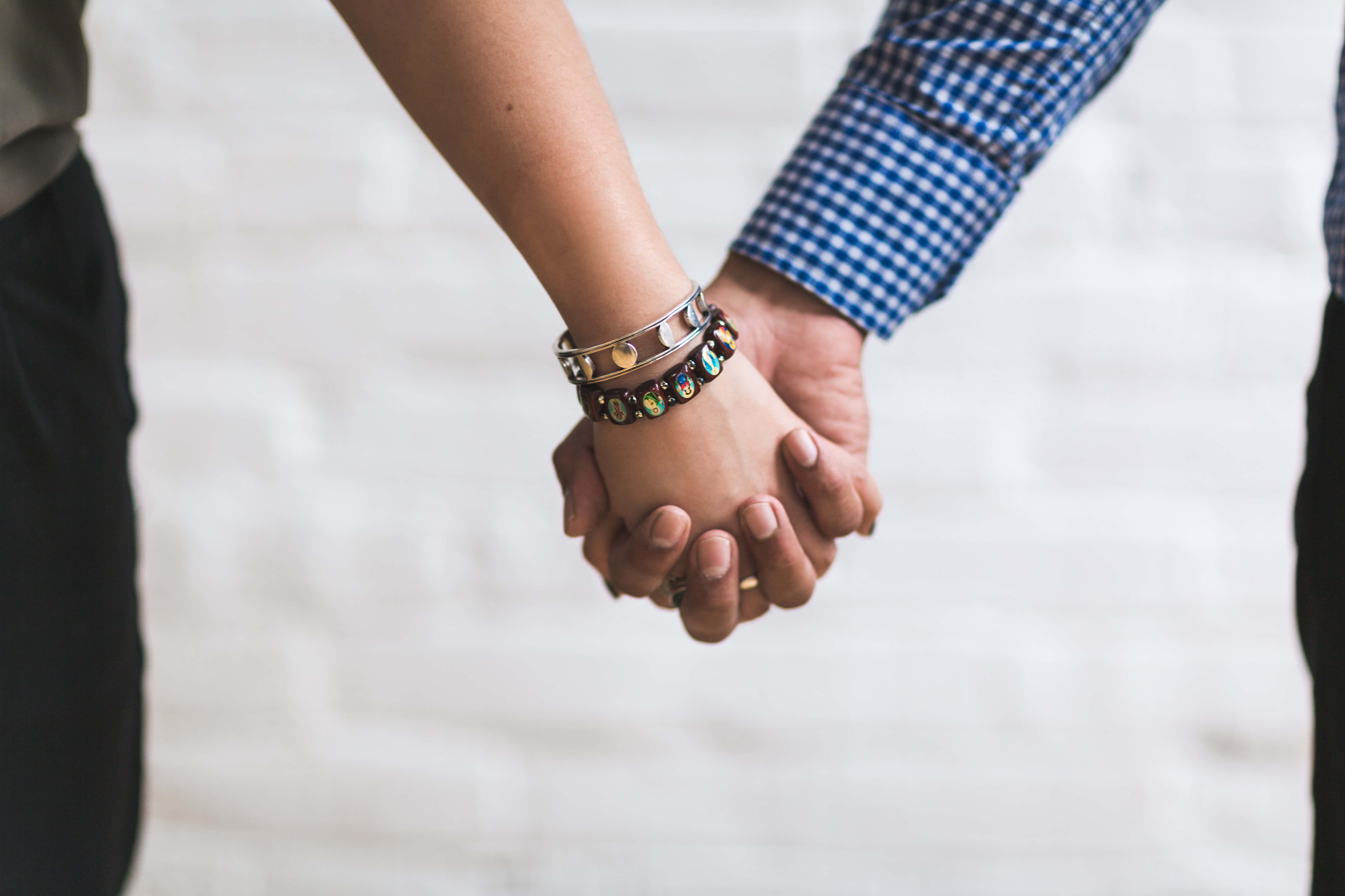Couple Holding Hand 桌布 壁紙 — 兩個人在白色磚牆前緊緊握住手，其中一人穿著藍色格紋襯衫和彩色珠珠手鍊。