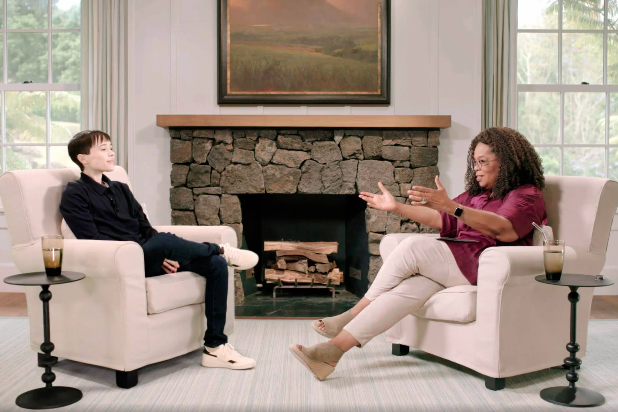 Oprah 桌布 壁紙 — 兩個人在石壁爐前的舒適扶手椅上交談，壁爐裡堆著木柴，上方掛著一幅風景畫。