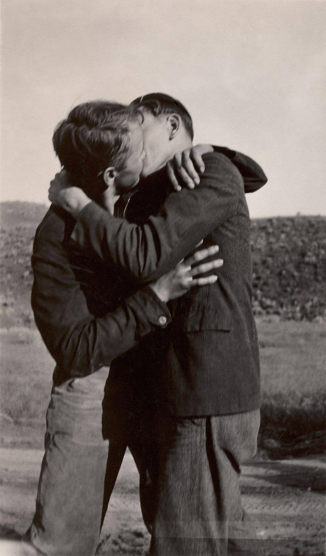 gay kiss 桌布 壁紙 — 兩個人在蒼白的天空下，在荒蕪的戶外景觀中熱情擁抱。