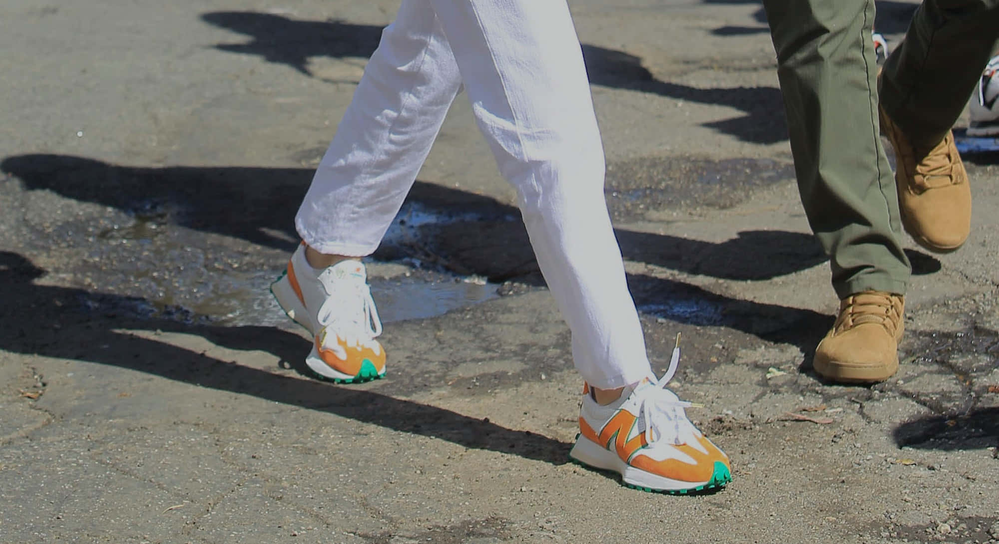 New Balance 桌布 壁紙 — 兩個人在裂開的人行道上行走的特寫，一個人穿著白色褲子和橙色運動鞋，另一個人穿著橄欖色褲子和棕褐色靴子。
