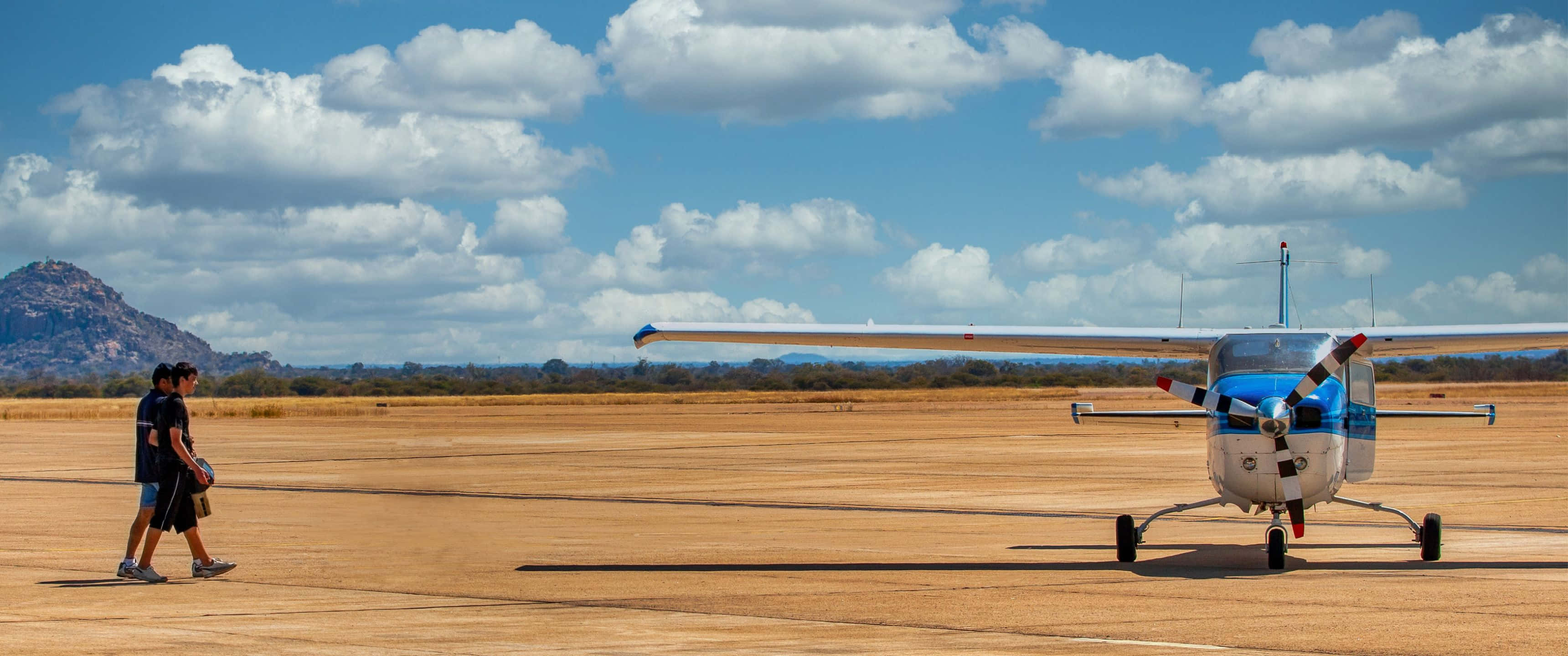 3440x1440p Small Planes 桌布 壁紙 — 兩個人在陽光明媚的沙漠跑道上，朝著一架藍白相間的小型螺旋槳飛機走去，天空湛藍，白雲朵朵。