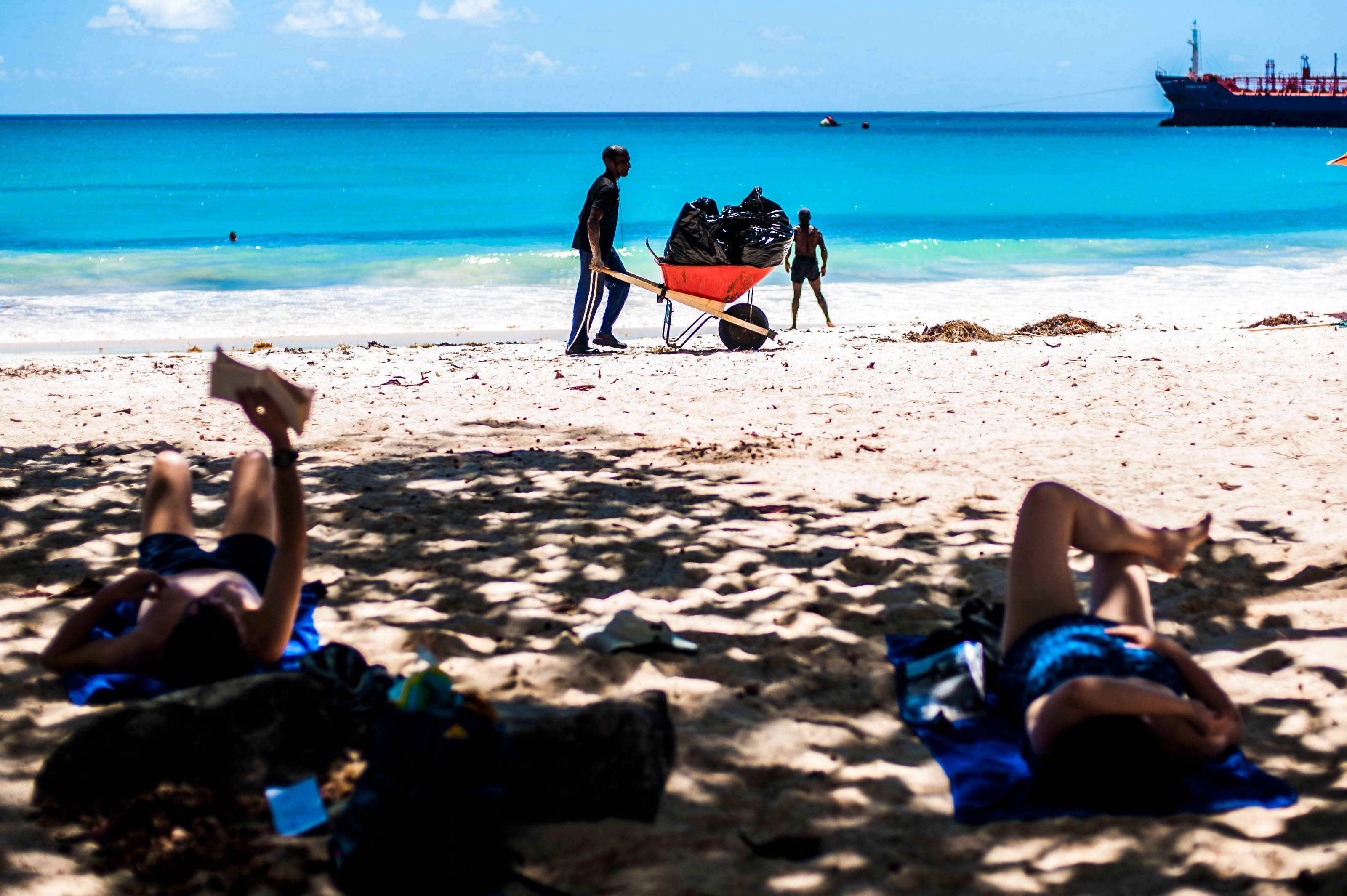 Barbados 桌布 壁紙 — 兩個人在陽光明媚的海灘上放鬆，而工人們用紅色手推車在碧綠海水附近運送垃圾。