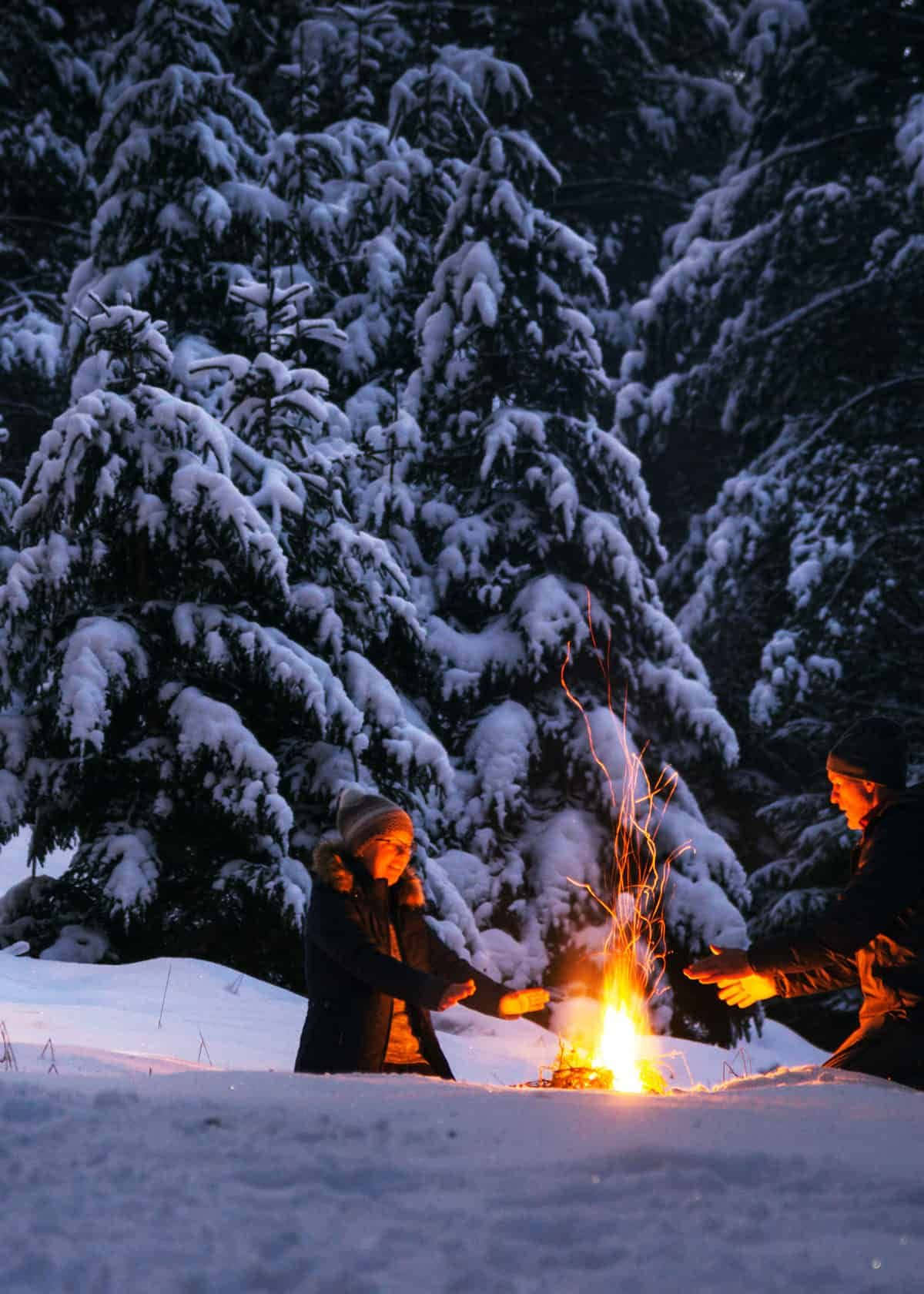 Winter Camping 桌布 壁紙 — 兩個人在黃昏時分的雪地森林中，圍著小營火取暖雙手。