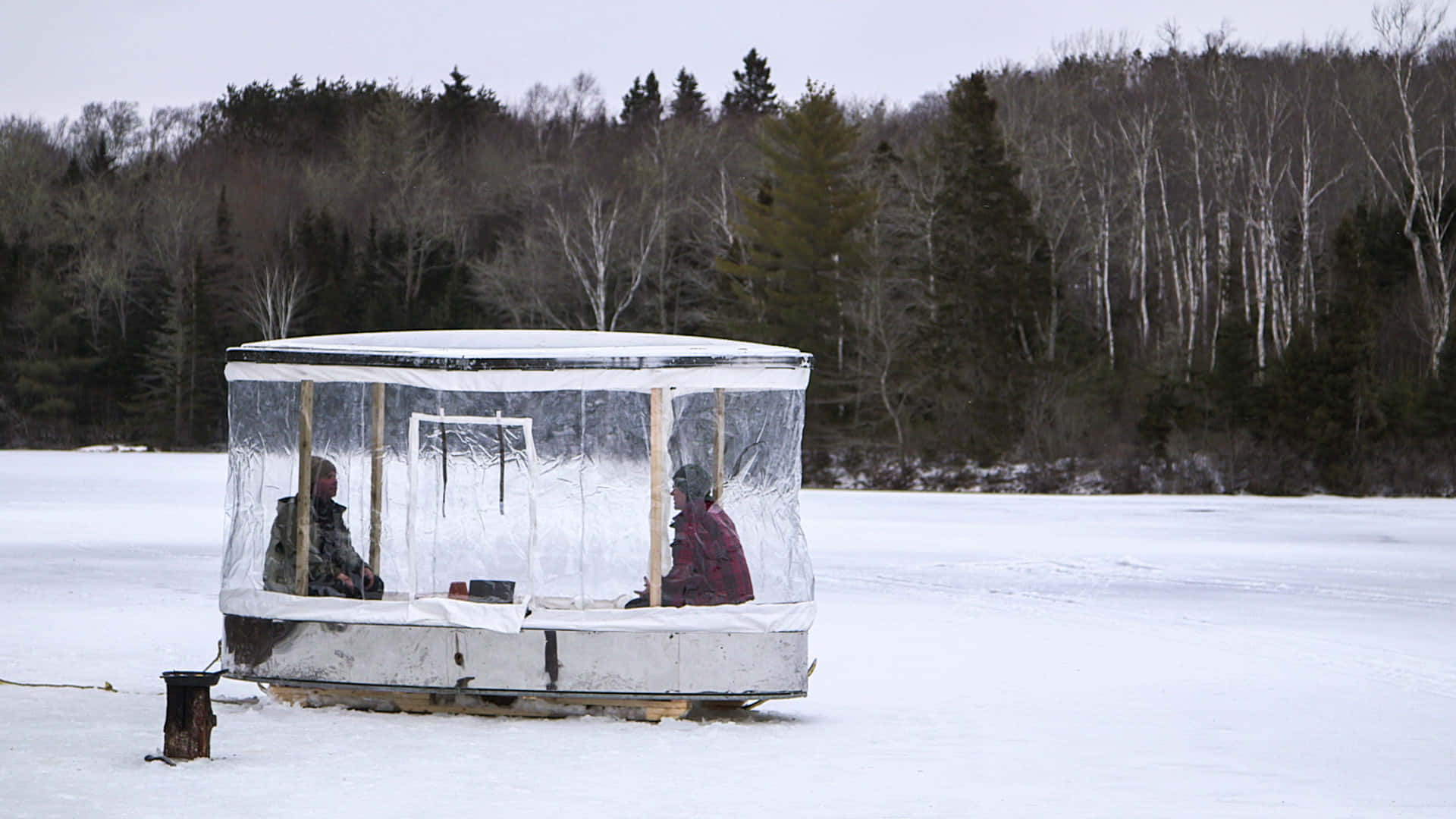 Ice Fishing 桌布 壁紙 — 兩個人坐在一個被雪覆蓋的冰釣棚內，位於一個被冬日禿樹環繞的冰凍湖面上。