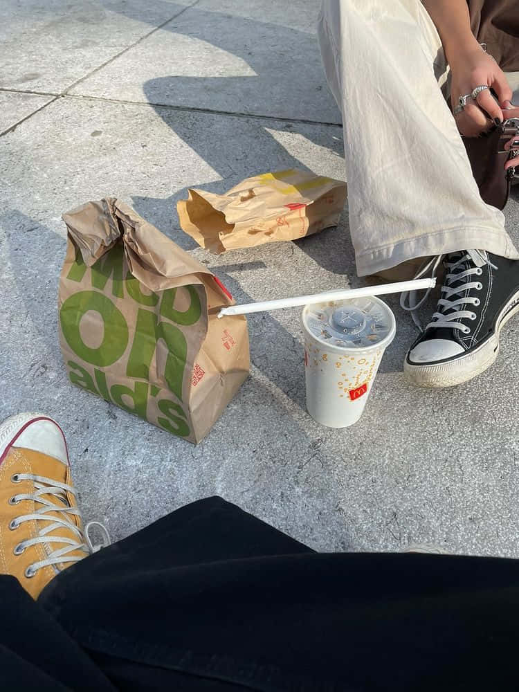 drinks 桌布 壁紙 — 兩個人坐在人行道上享受麥當勞的食物和飲料，旁邊有運動鞋和紙袋。
