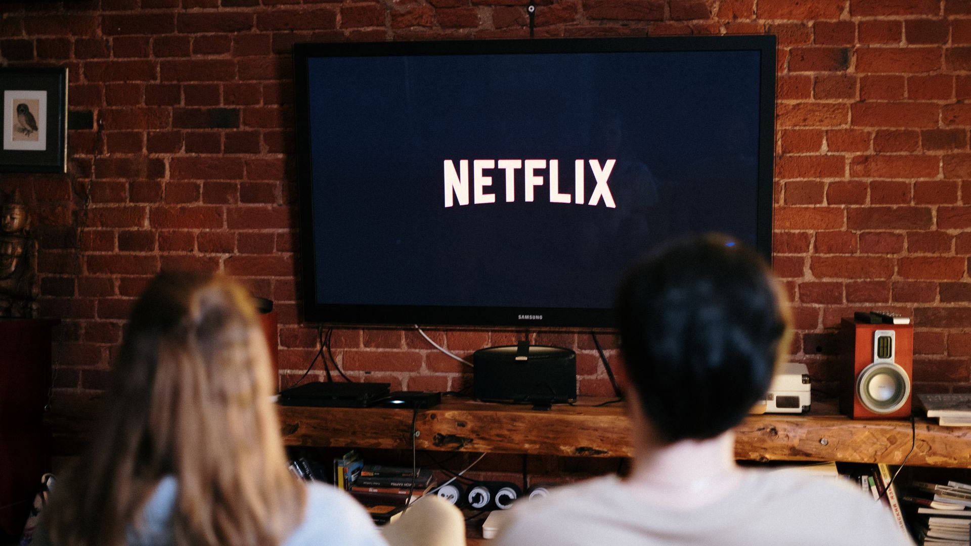 Netflix 桌布 壁紙 — 兩個人坐在沙發上，觀看安裝在紅磚牆上的電視播放Netflix，身處舒適的客廳。
