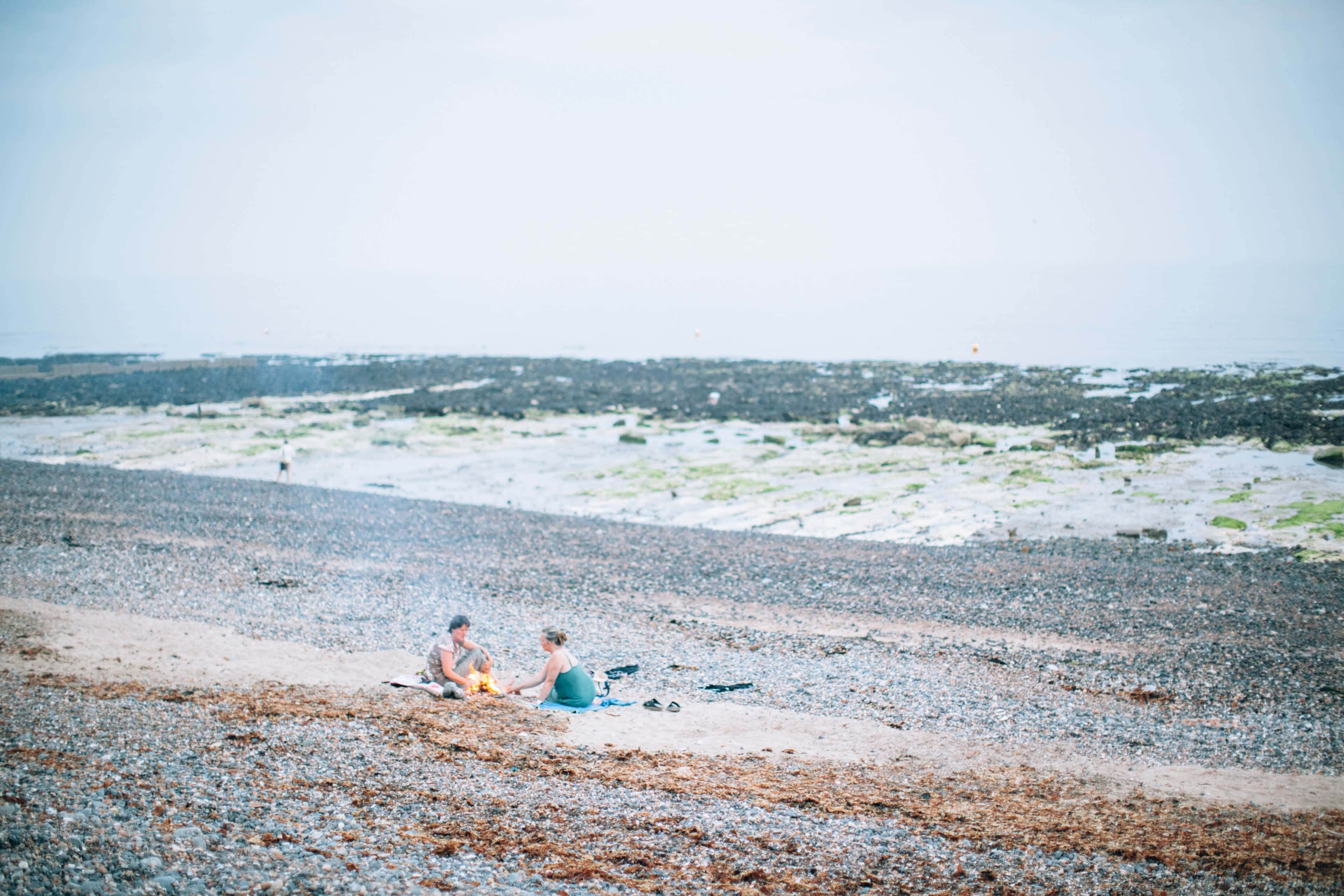Beach Picnic 桌布 壁紙 — 兩個人坐在黃昏的鵝卵石海灘上，照料著一個小營火，周圍散落著海藻。