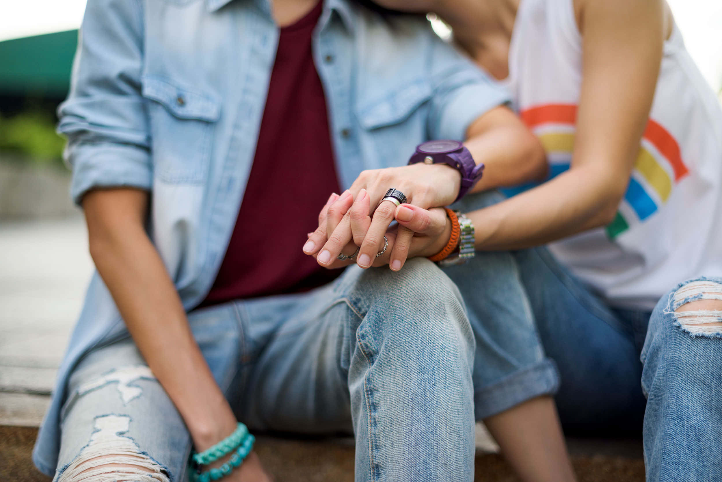 Holding Hands 桌布 壁紙 — 兩個人坐得很近，手牽手，其中一人的襯衫上有彩虹驕傲旗幟，穿著休閒牛仔服裝。