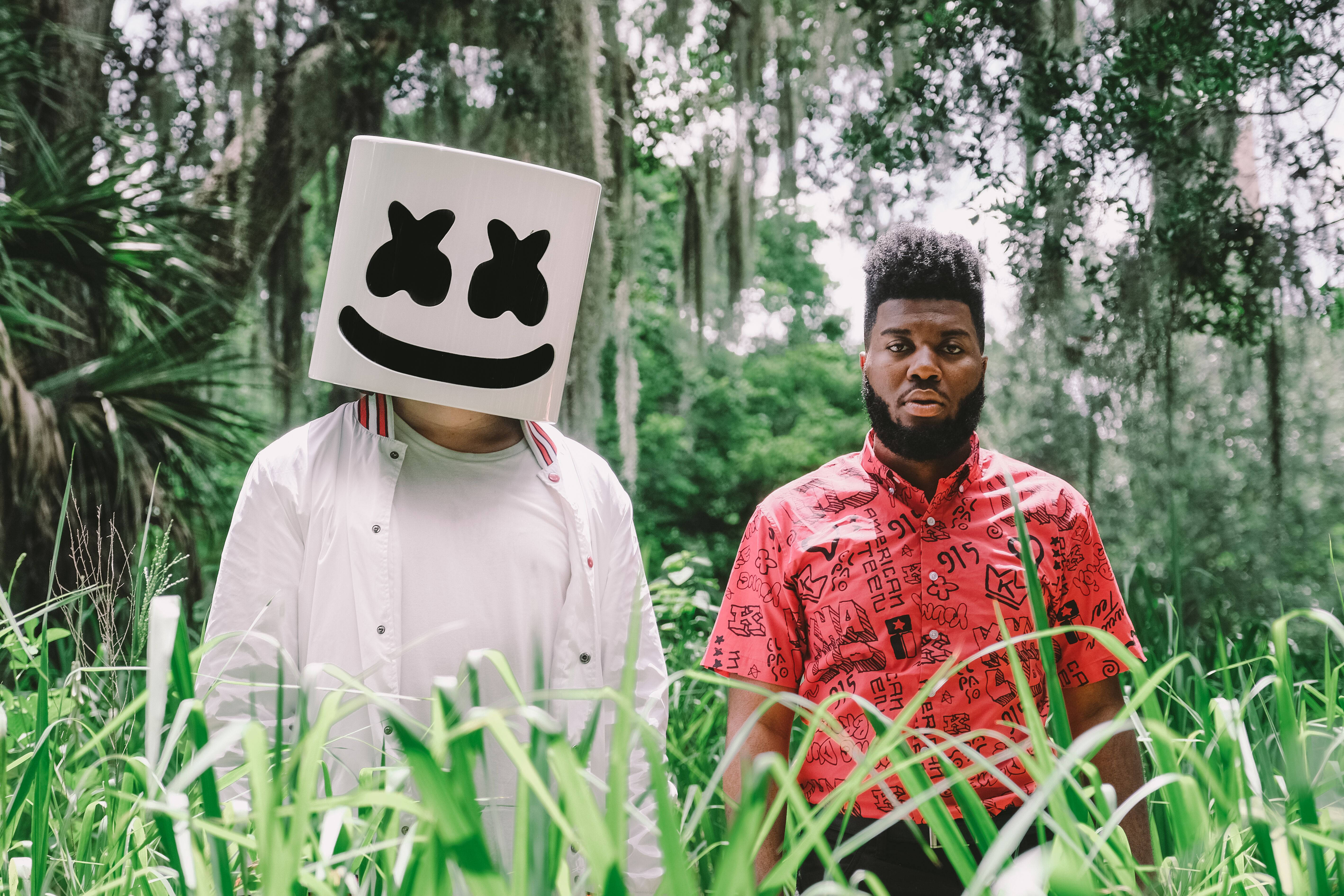 Marshmello 4K 桌布 壁紙 — 兩個人影站在茂密的沼澤中，一人戴著白色棉花糖面具，另一人穿著紅色圖案襯衫，周圍環繞著綠色樹葉和垂掛的苔蘚。