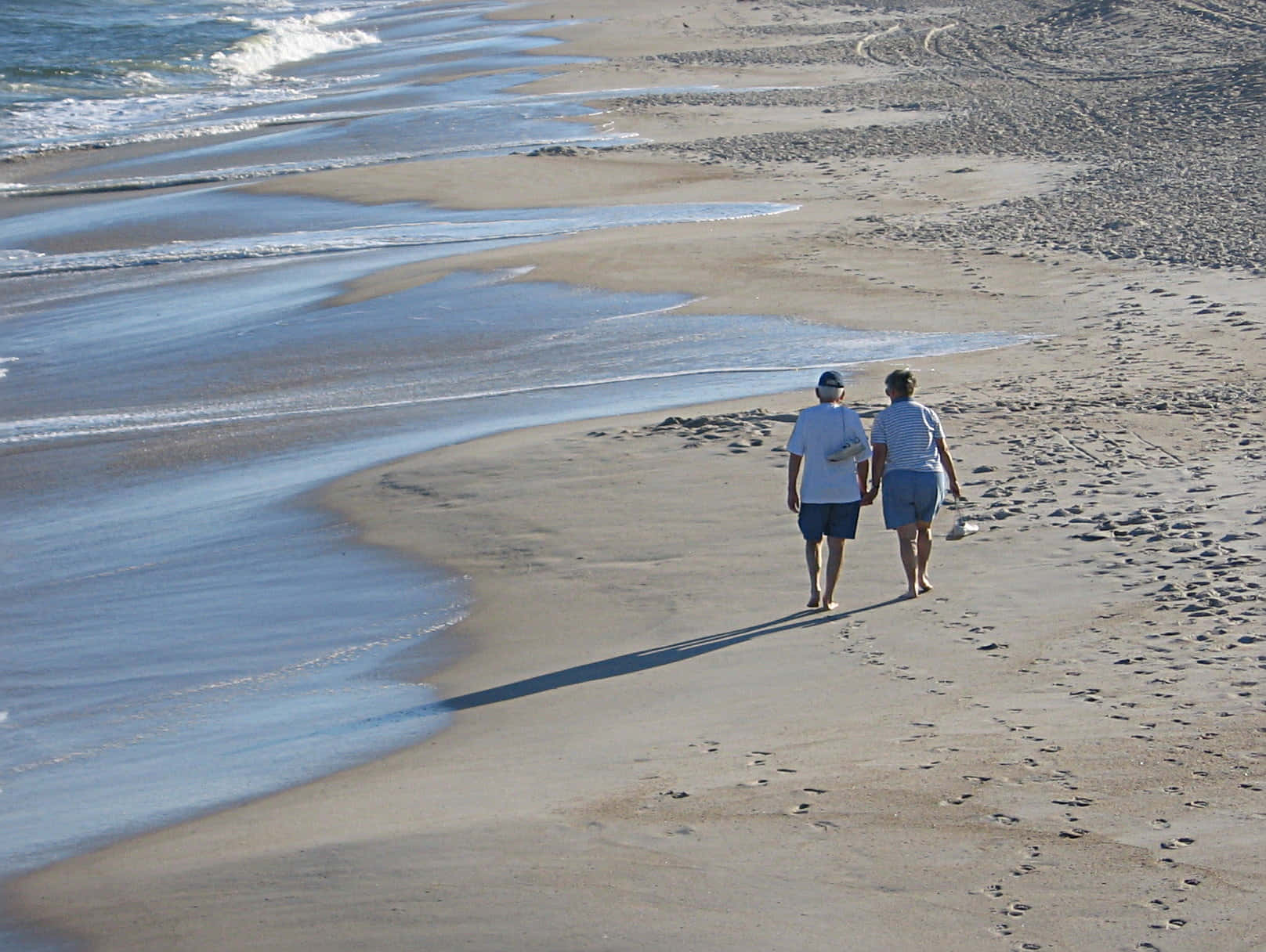 Beach Walk 桌布 壁紙 — 兩個人手牽手漫步在陽光明媚的海灘上，溫柔的海浪拍打著海岸。