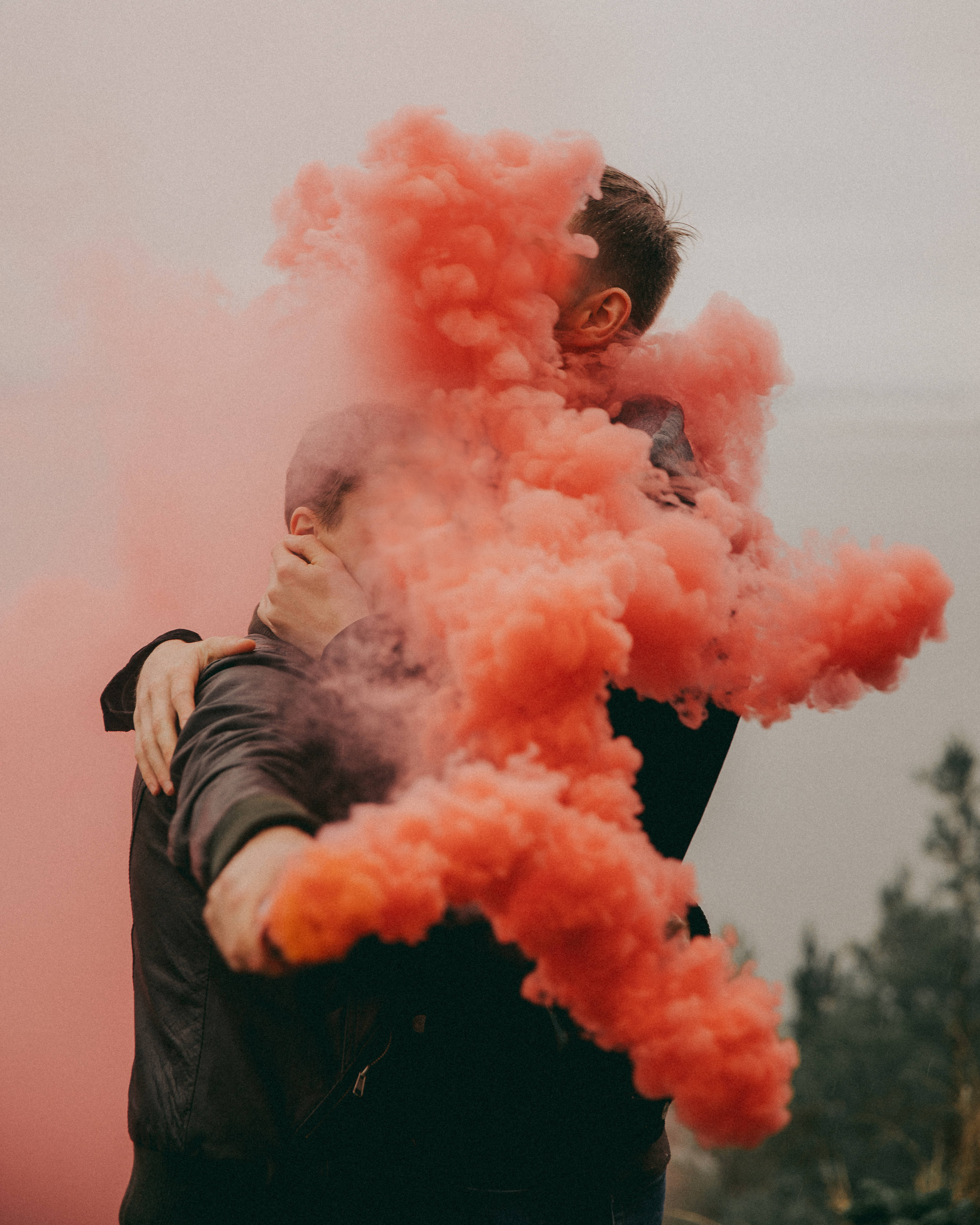 men kissing 桌布 壁紙 — 兩個人擁抱，鮮豔的珊瑚色煙霧在他們周圍翻騰，背景是柔和的戶外環境。