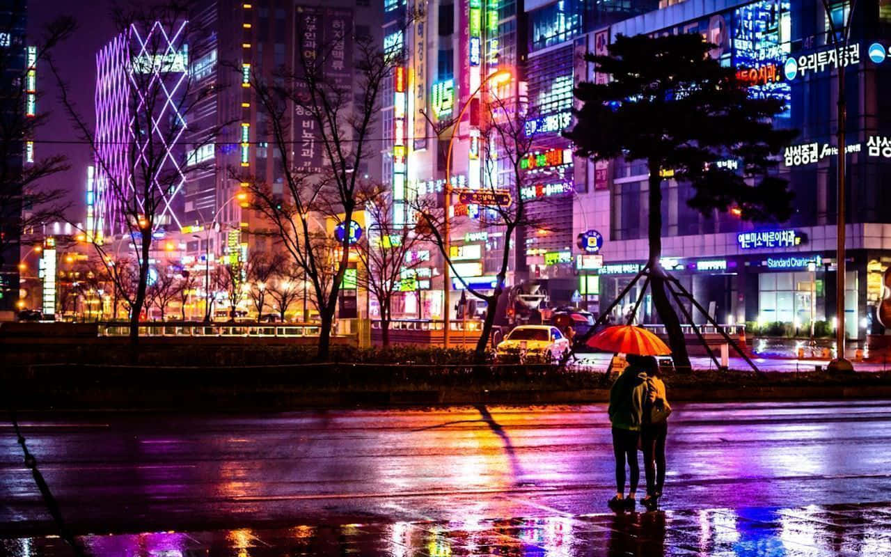 Aesthetic Korean 桌布 壁紙 — 兩個人物在橙色雨傘下走過雨濕的街道，街道反射著夜間高聳城市建築的霓虹燈光。