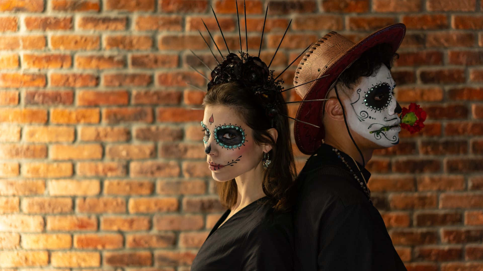 Horror Costumes 桌布 壁紙 — 兩個人畫著精緻的糖骷髏妝容，背靠背站在溫暖的磚牆前，其中一人手持一朵紅花。
