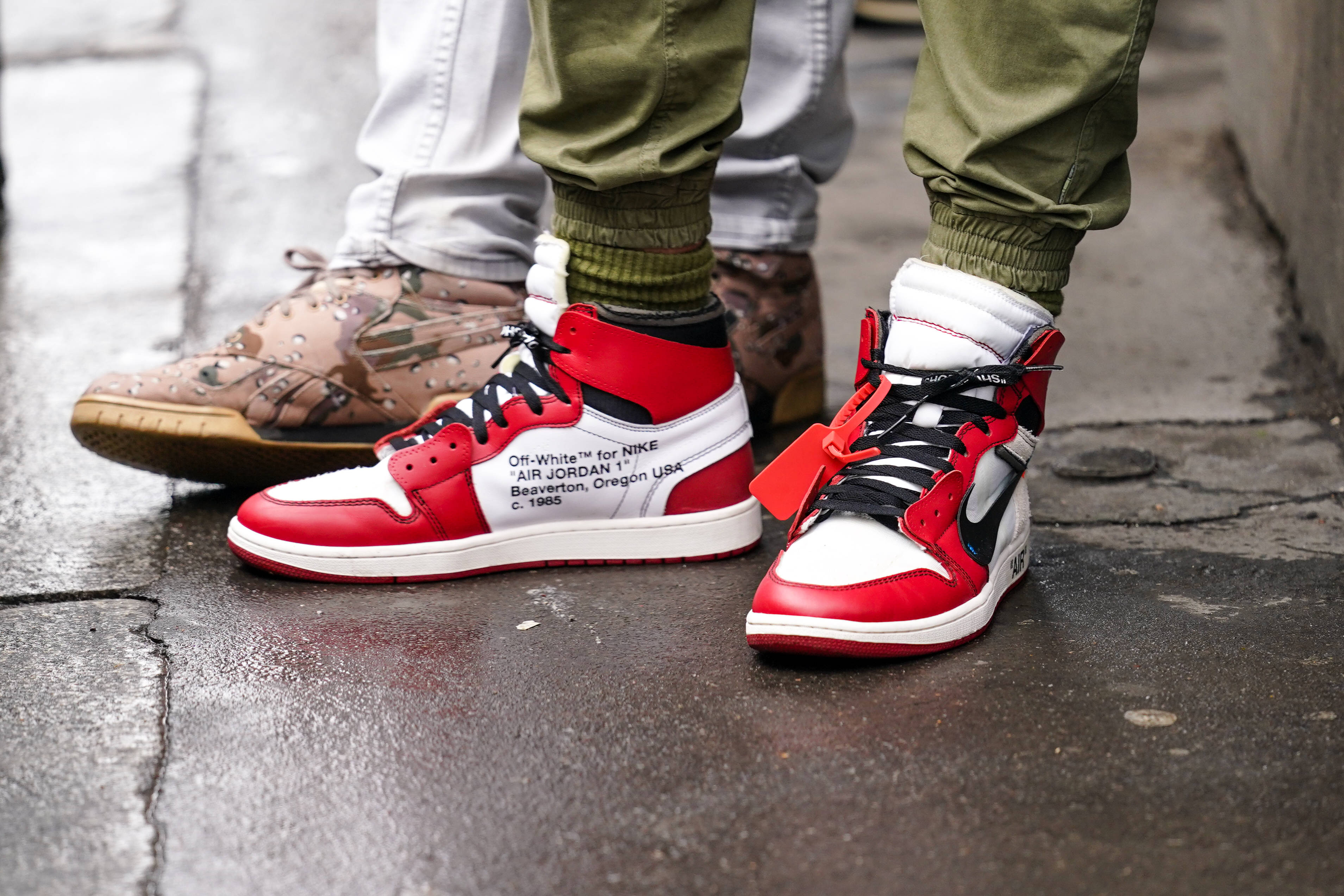 Nike Air Jordan 1 桌布 壁紙 — 兩個人的腳近距離特寫，穿著紅、白、黑色運動鞋在濕的城市人行道上。