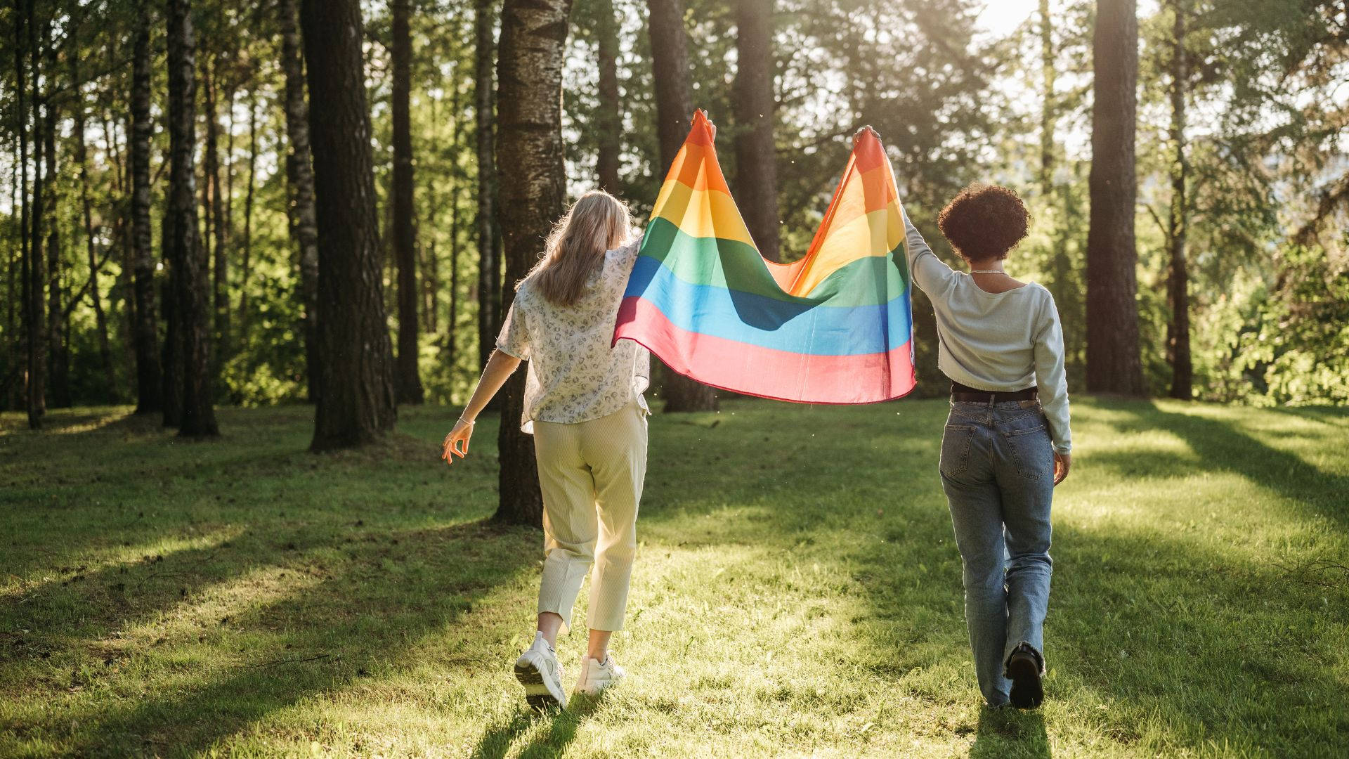 Subtle Lgbt 桌布 壁紙 — 兩個人穿過陽光明媚的森林，高舉著鮮豔的彩虹旗。