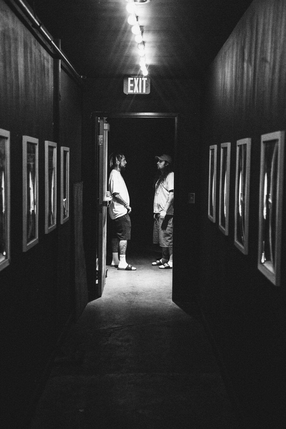 suicideboys 桌布 壁紙 — 兩個人站在一個昏暗的走廊裡，走廊兩旁掛著裱框的圖片，僅由一盞天花燈和一個出口標誌照亮。