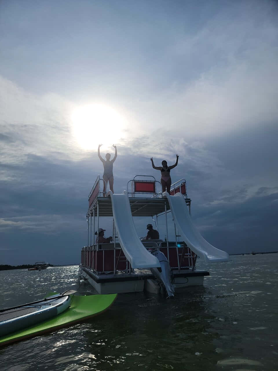 Crab Island 桌布 壁紙 — 兩個人站在漂浮的滑水梯平台上，舉起雙臂，映襯著明亮的太陽和多雲的天空。