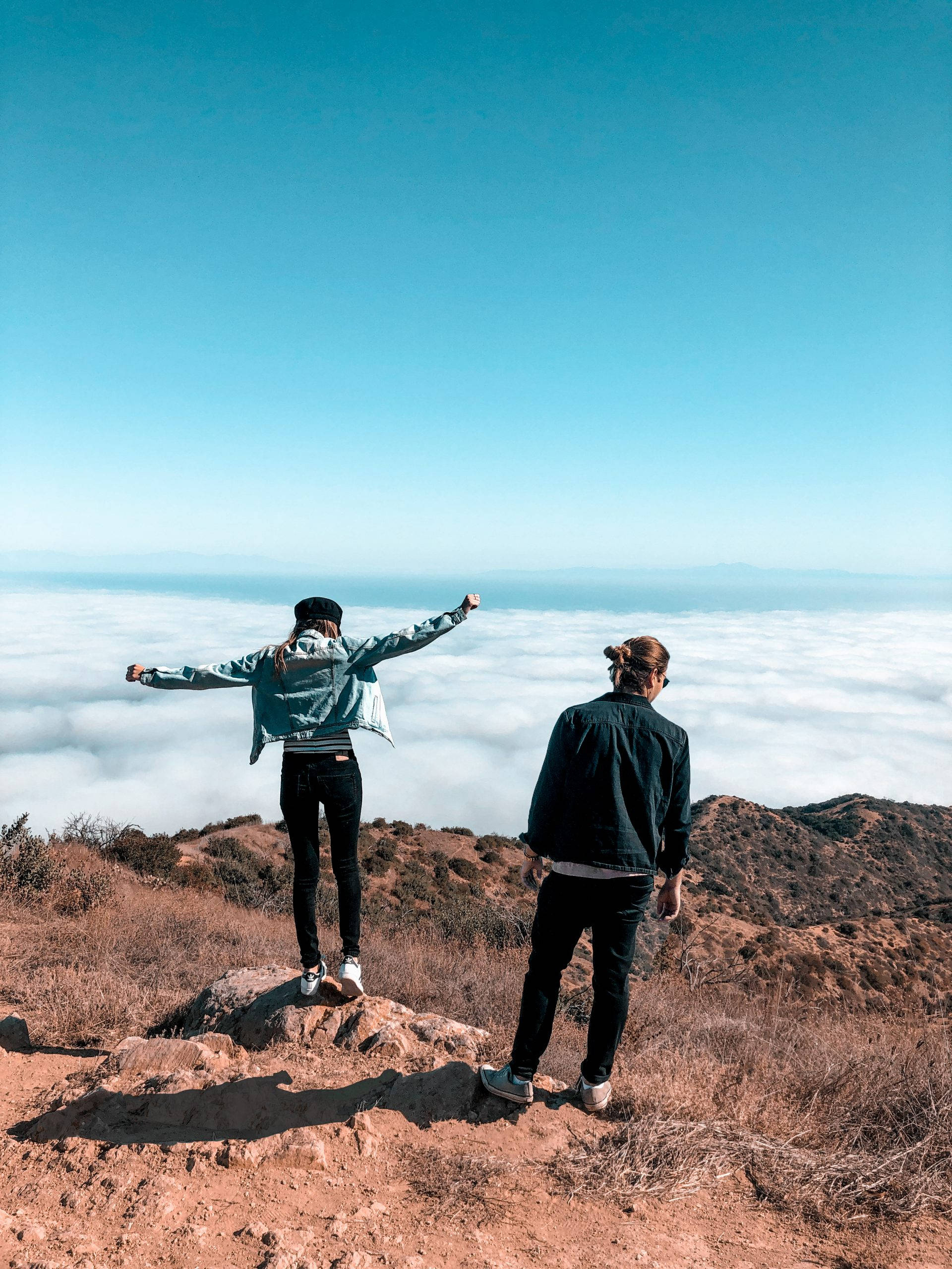 Hiking 桌布 壁紙 — 兩個人站在陽光明媚的山頂上，俯瞰著晴朗藍天下廣闊的雲海。