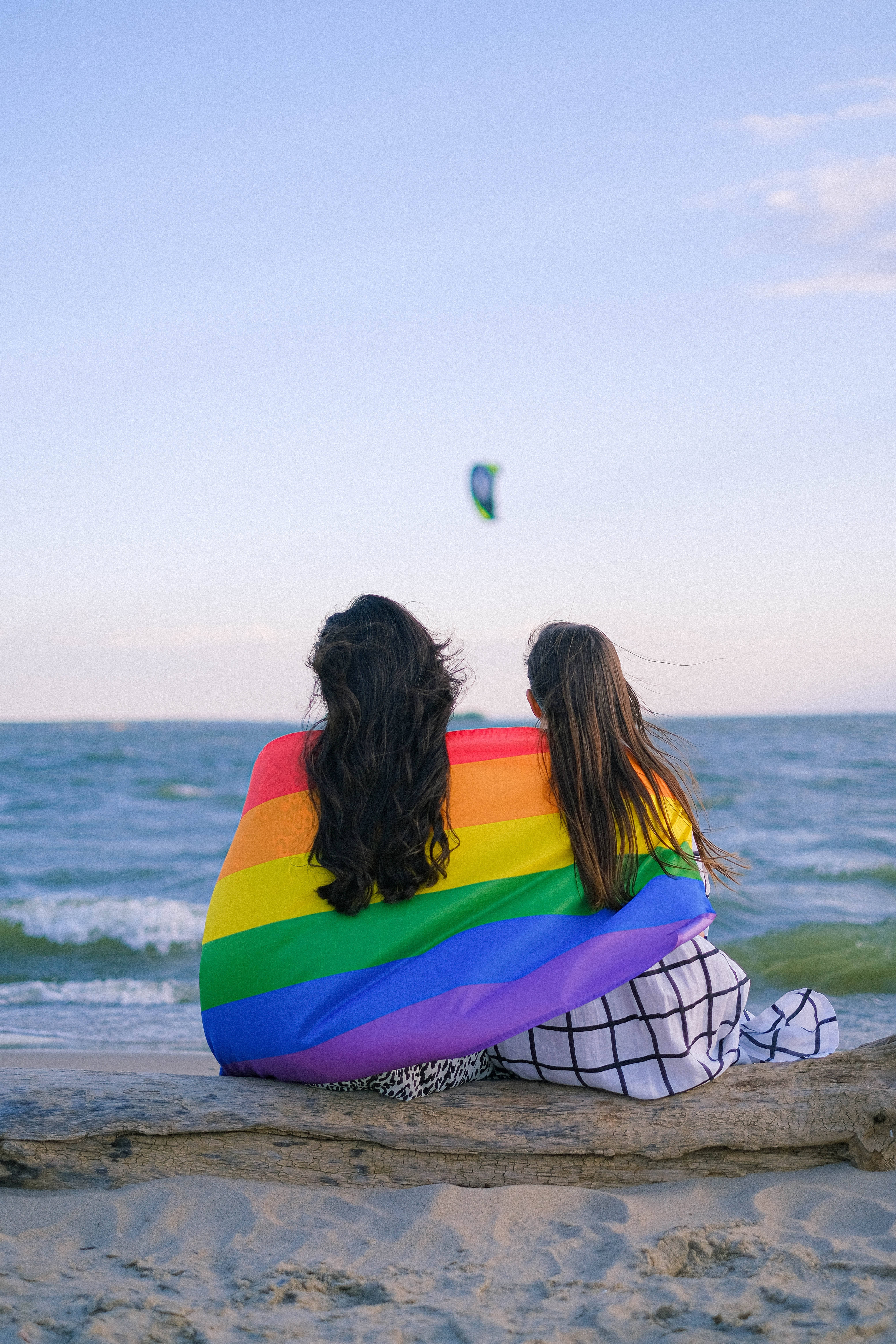 Pride Flag 桌布 壁紙 — 兩個人背對背坐在沙灘木頭上，裹著彩虹旗，看著風箏在日落時分在海洋上空航行。