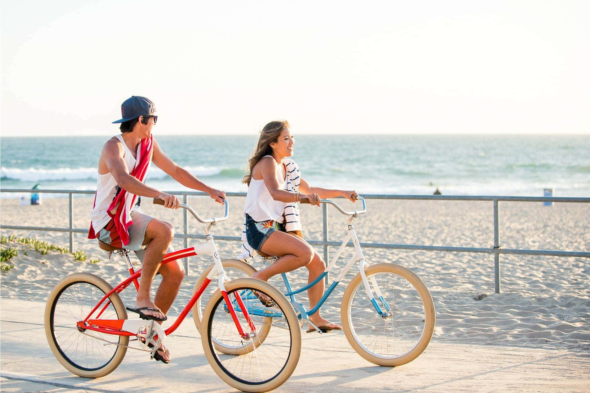 Beach Cruiser 桌布 壁紙 — 兩個人騎著色彩繽紛的沙灘巡航車沿著海濱長廊行駛，身後是海浪和明亮的天空。