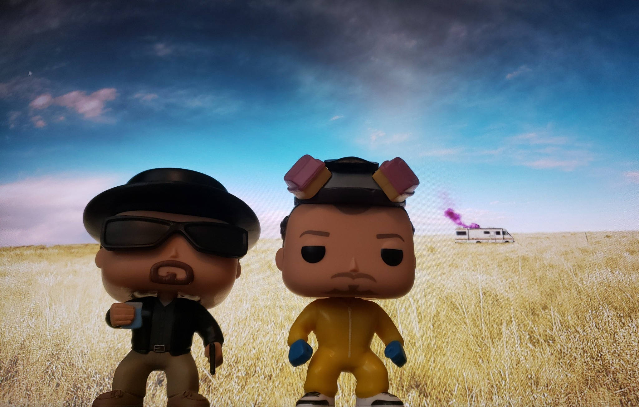 pop 桌布 壁紙 — 兩個來自《絕命毒師》的Funko Pop公仔站在乾燥的草地上，天空戲劇性，遠處有一輛露營車。