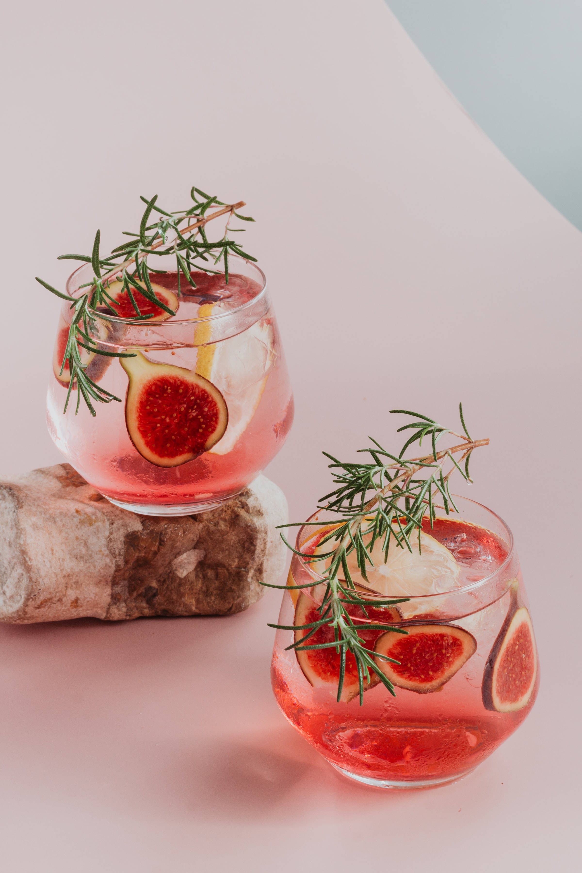 drinks 桌布 壁紙 — 兩個優雅的玻璃杯裝滿粉紅色飲料，飾以無花果片、迷迭香和檸檬，置於柔和粉彩背景上。