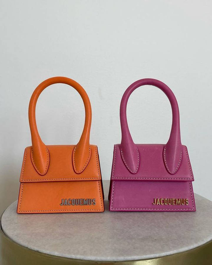 Handbag 桌布 壁紙 — 兩個充滿活力的橙色和洋紅色Jacquemus迷你手提包，帶有彎曲手柄，並排放在大理石底座上，背景是純白牆壁。