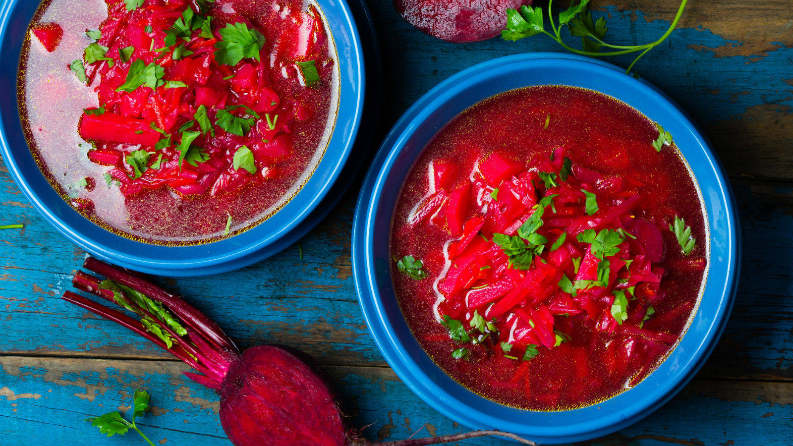 beetroot 桌布 壁紙 — 兩個充滿活力的藍色碗，盛滿濃郁的紅色羅宋湯，上面撒上新鮮歐芹，放在一個質樸、風化的藍色木桌上。