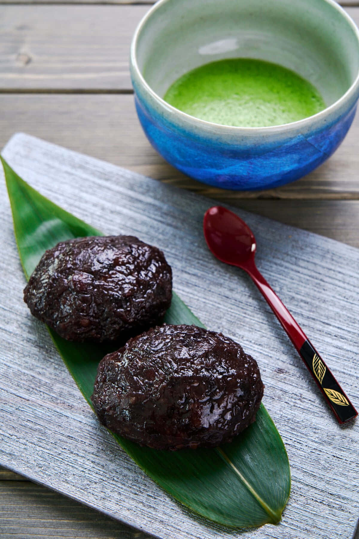 Japanese Sweets 桌布 壁紙 — 兩個光滑的深色麻糬放在綠色竹葉上，旁邊有一個藍色碗裝著鮮綠抹茶和紅色湯匙，置於風化木上。